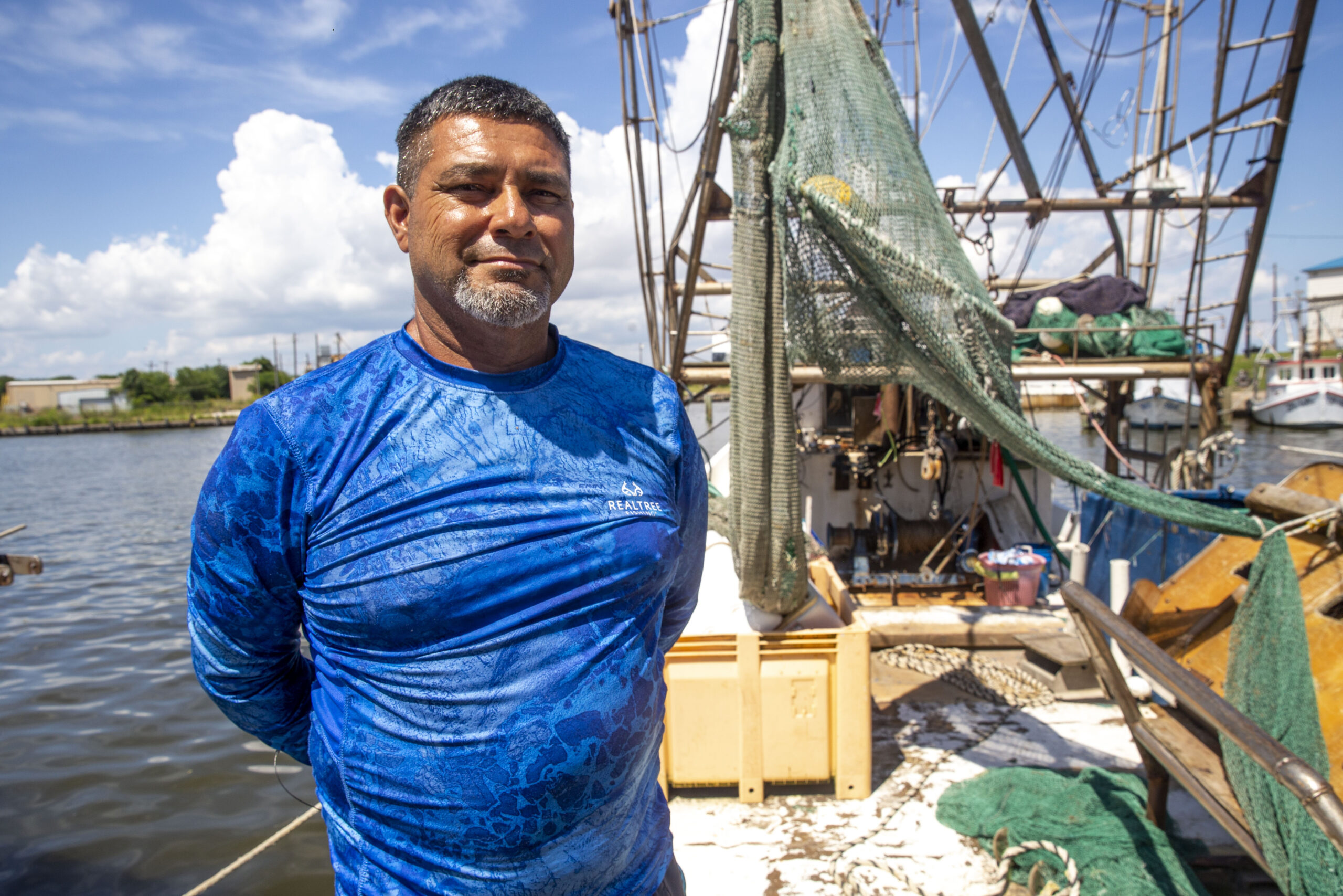 Maurico Blanco is a Latino man posing in a blue shirt, proudly showing off the rigging of his shirmping boat with the waters of Lavaca Bay, photographed under a partly cloudy sky, behind him.
