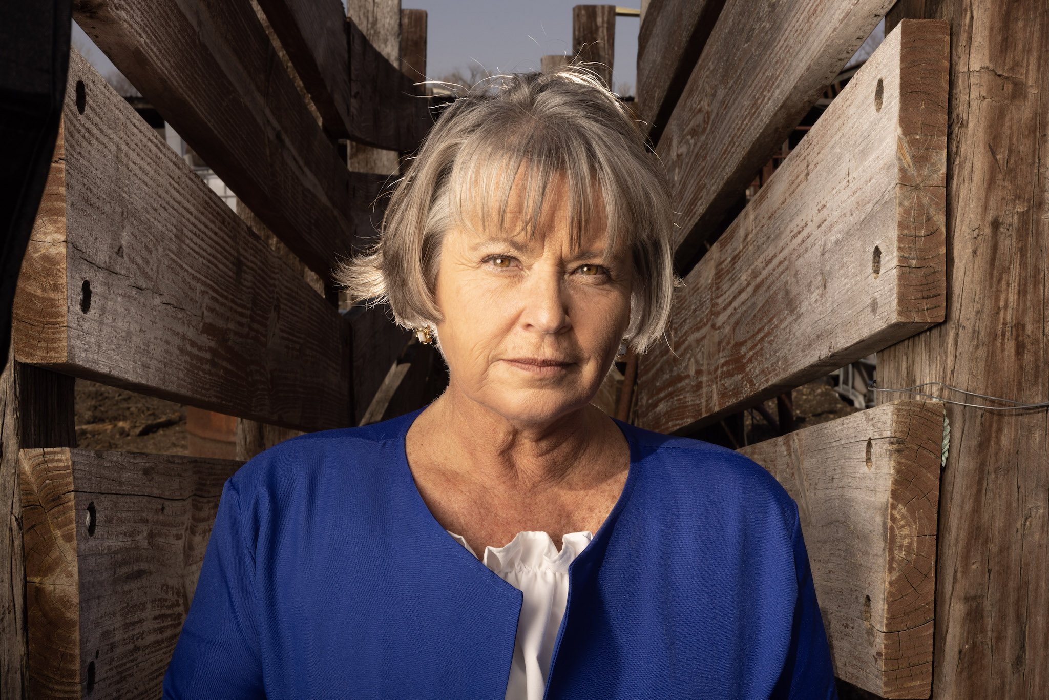 Mary Kelleher is a gray-haired white woman, wearing a blue jacket over a white blouse. She is photographed outdoors in a barn.
