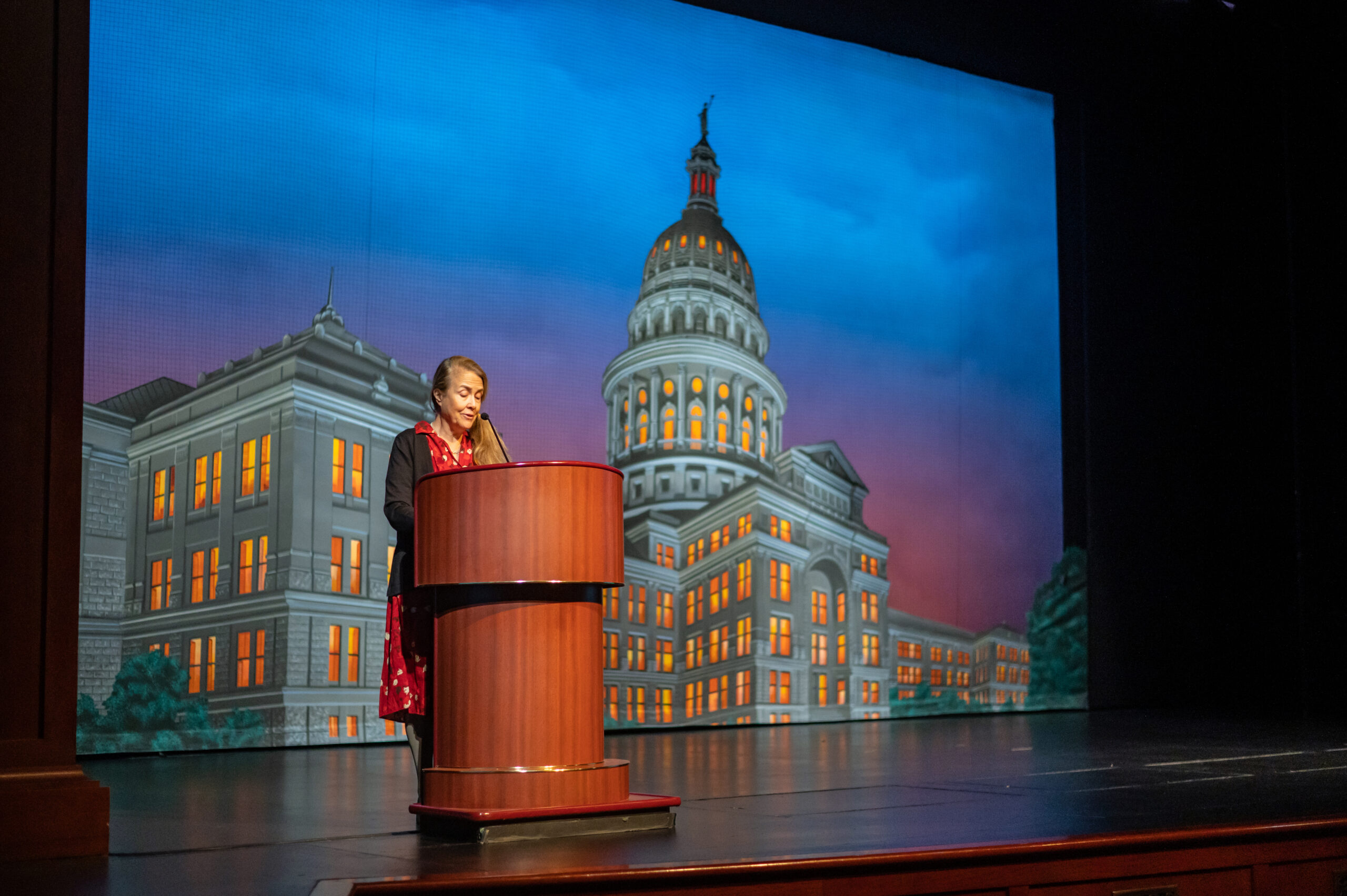 Naomi Shihab Nye, wearing a polka dot blouse under a blue suit jacket, speaks at a podium in front of a projection of the Texas Capitol building.