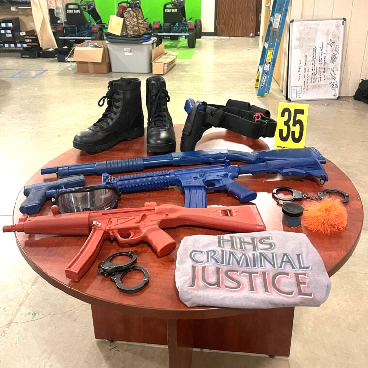 A classroom in a table covered with real and mock prison gear including boots, pretend rifles, and a "HHS Criminal Justice" shirt.