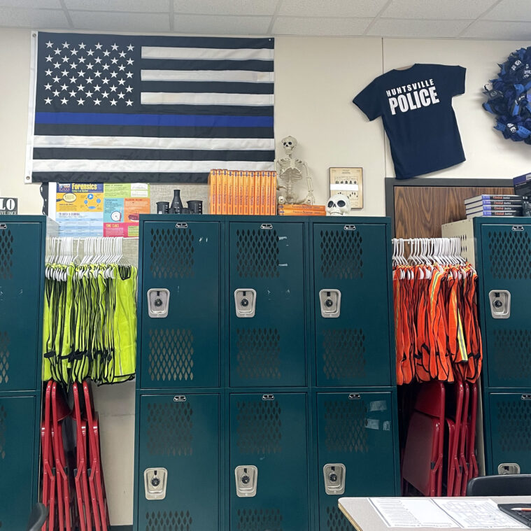 Vests and lockers line the walls, along with a thin blue line flag and a Huntsville Police t-shirt.
