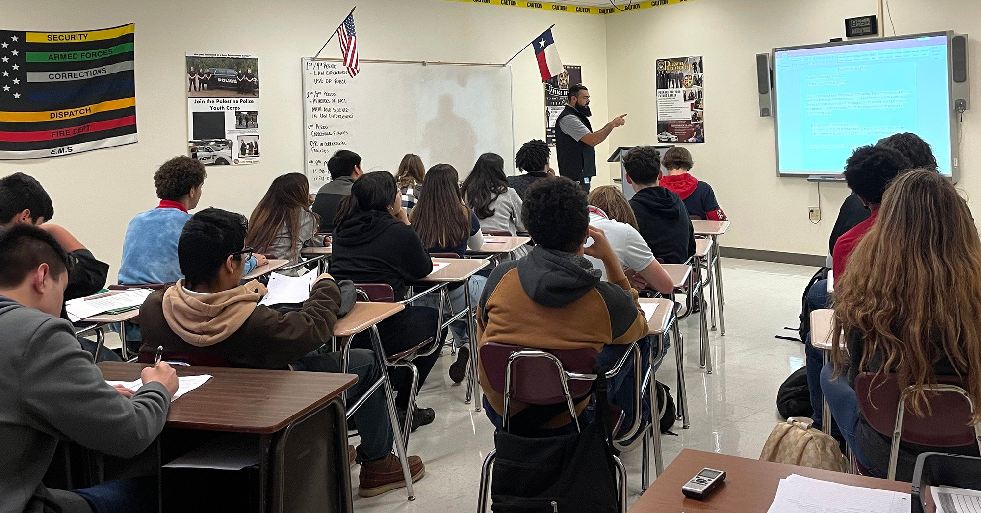 A teacher with a beard lectures a classroom of high school students about law enforcement and prison careers, in front of a powerpoint presentation. Pro-prison and law enforcement posters and a law enforcement flag cover the walls.
