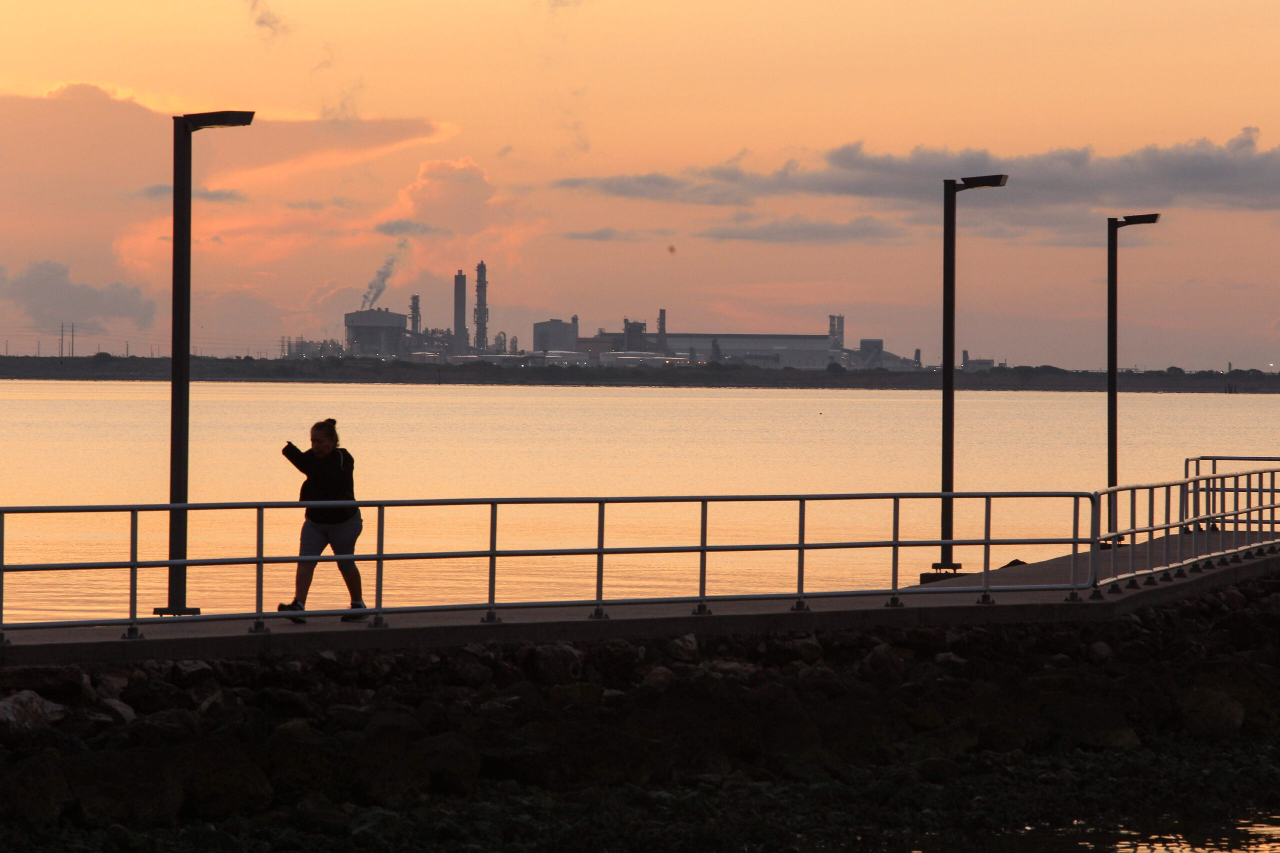 A person walks along a fenced in pier along the water, at sunset, with a steaming refinery/oil terminal across the Gulf.