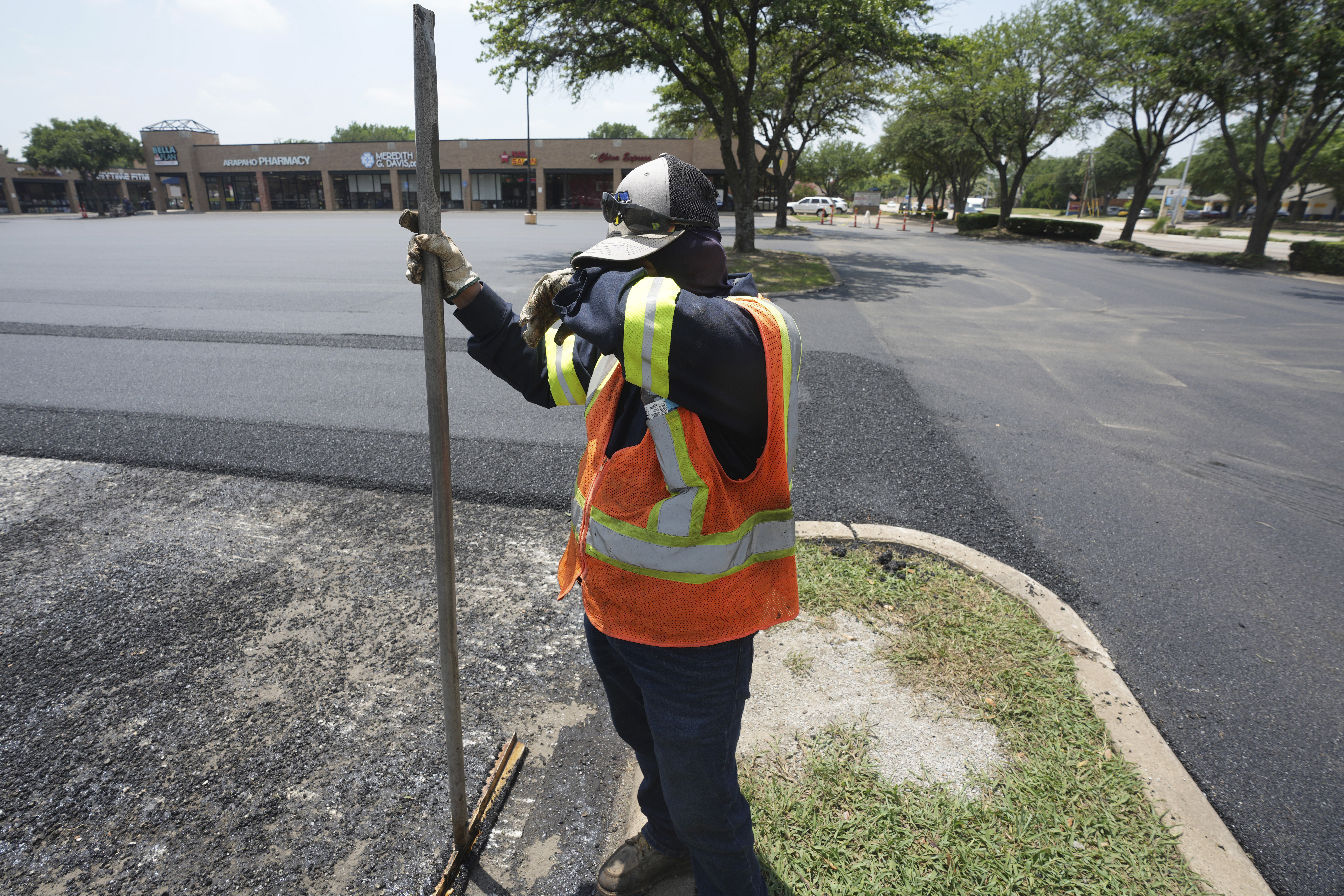 A man in a construction uniform including safety vest, pauses while shoveling asphalt in a parking lot, wiping his face with his arm to remove the excess sweat. The sun bakes down on the surface of the parking lot where he's working.