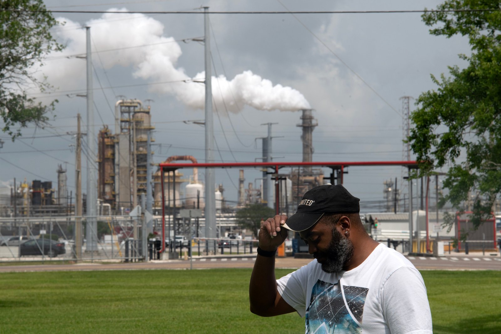 Christopher Jones, a Black man in a ball cap and t-shirt, poses with his head to the side and down, in front a steaming oil refinery.
