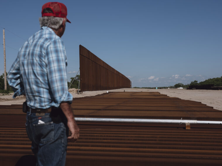 A man in a red mesh trucker cap, blue and white plaid shirt and blue jeans looks at border wall construction supplies along a desolate stretch of land.