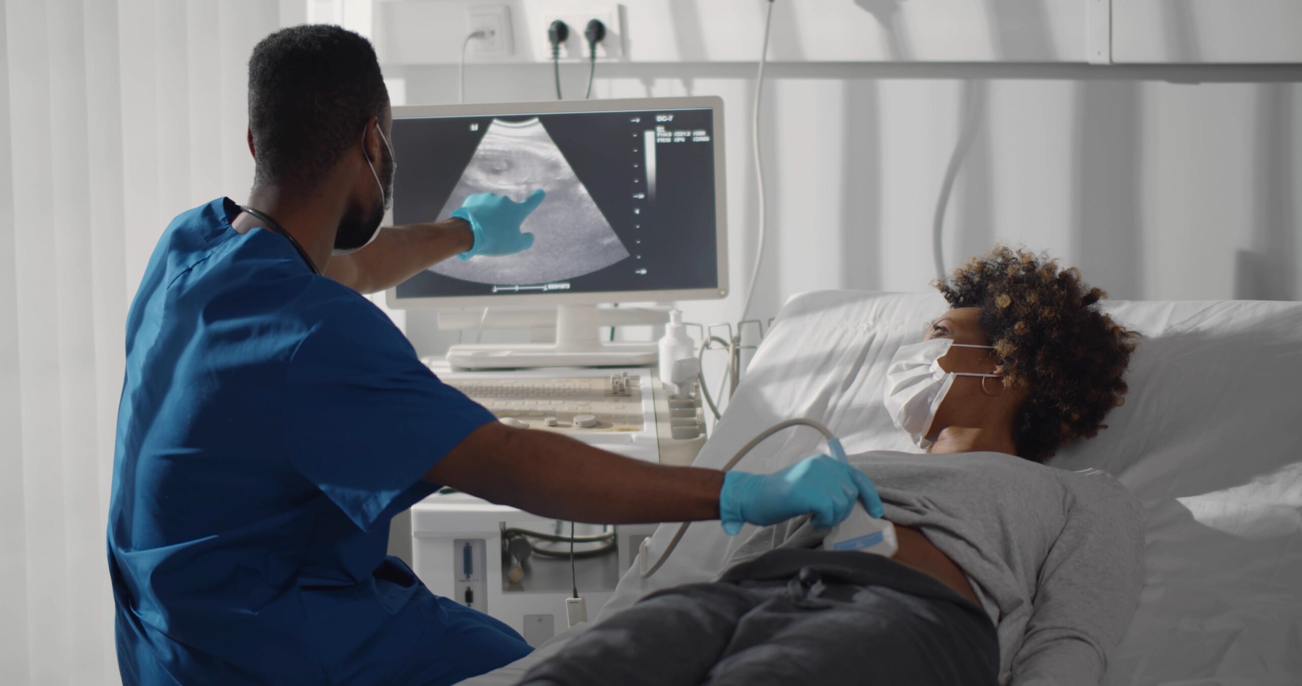 A masked Black woman laying in a hospital bed receives an ultrasound from a Black medical technician or doctor, who is pointing at the computer screen showing the infant.