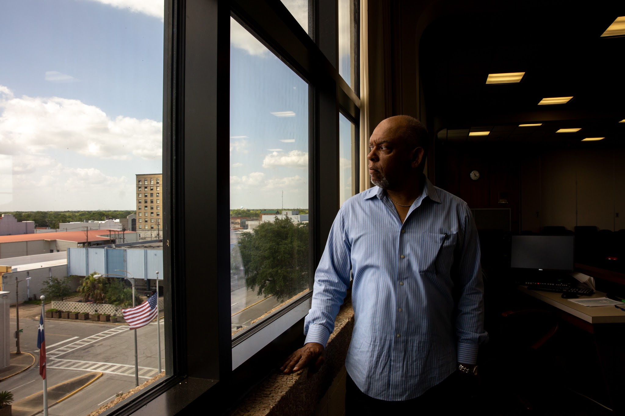 John Beard stands in a dimly lit office, looking out a grid of large glass windows onto a parking lot. he is wearing a button down shirt, and has a serious expression.