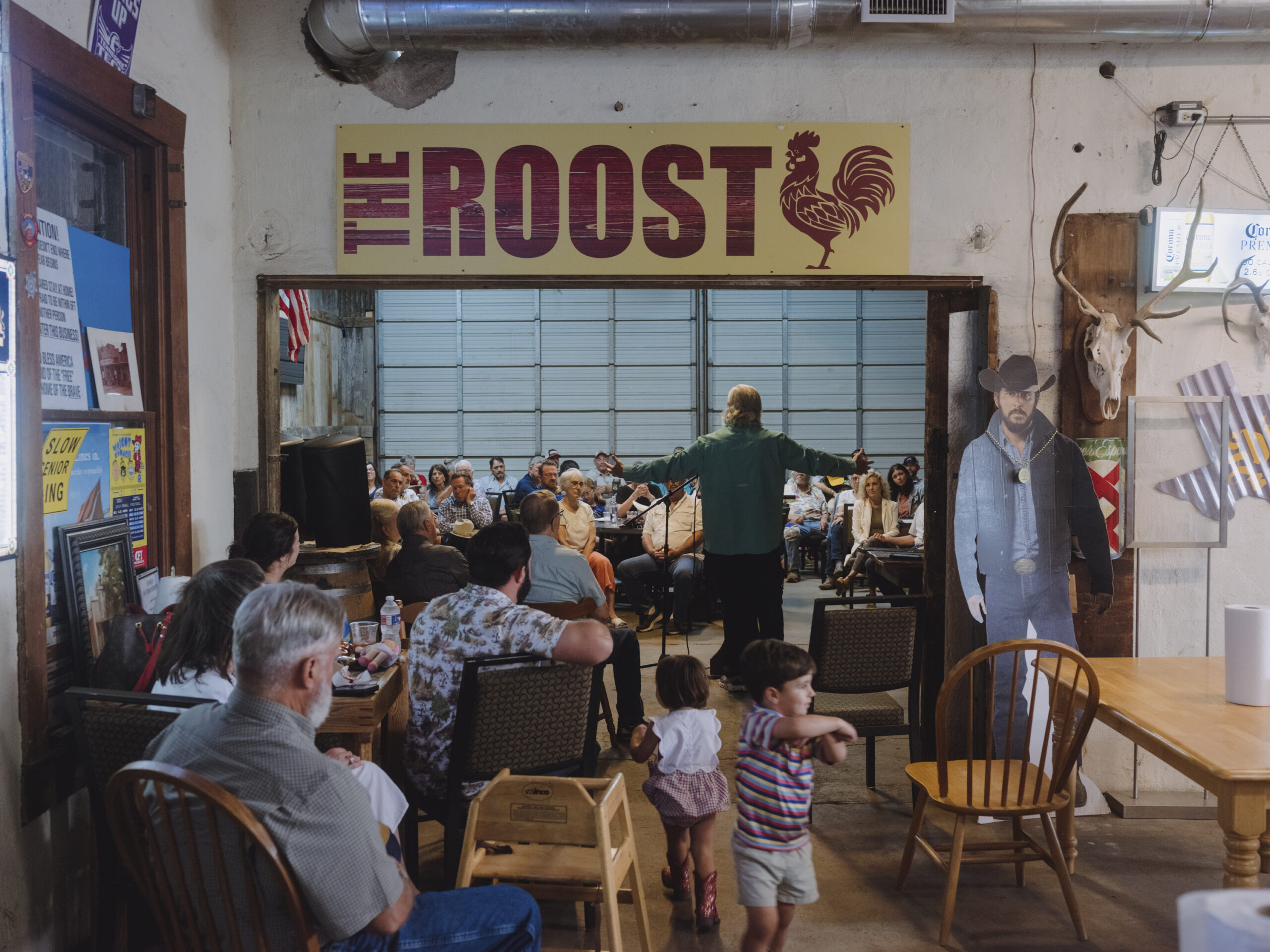 Kids run around the crowded main room of the general store, which is cluttered with knick-knacks. A preacher speaks at a microphone with arms outstretched, wearing casual clothing.