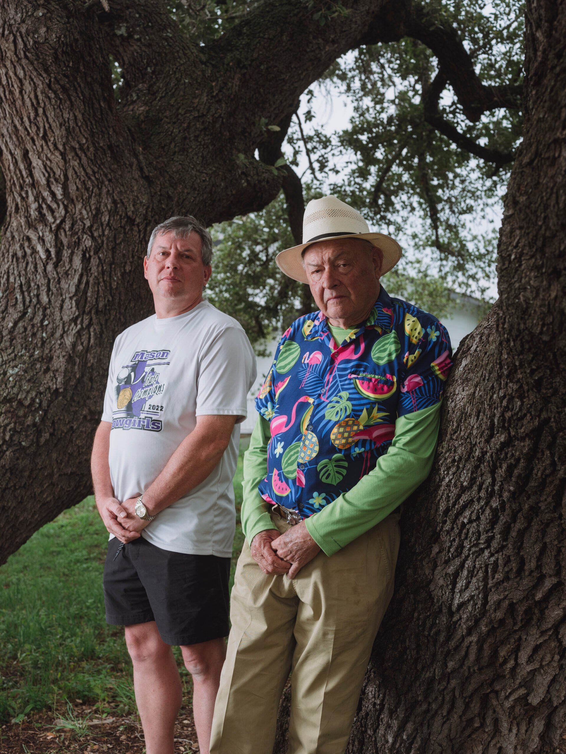Marc and Randy Leifeste, two middle aged white men, stand leaning against an ancient oak. Marc is in shorts and a t-shirt while Randy is wearing a Hawaiian shirt, a Panama hat, and tan khakis.