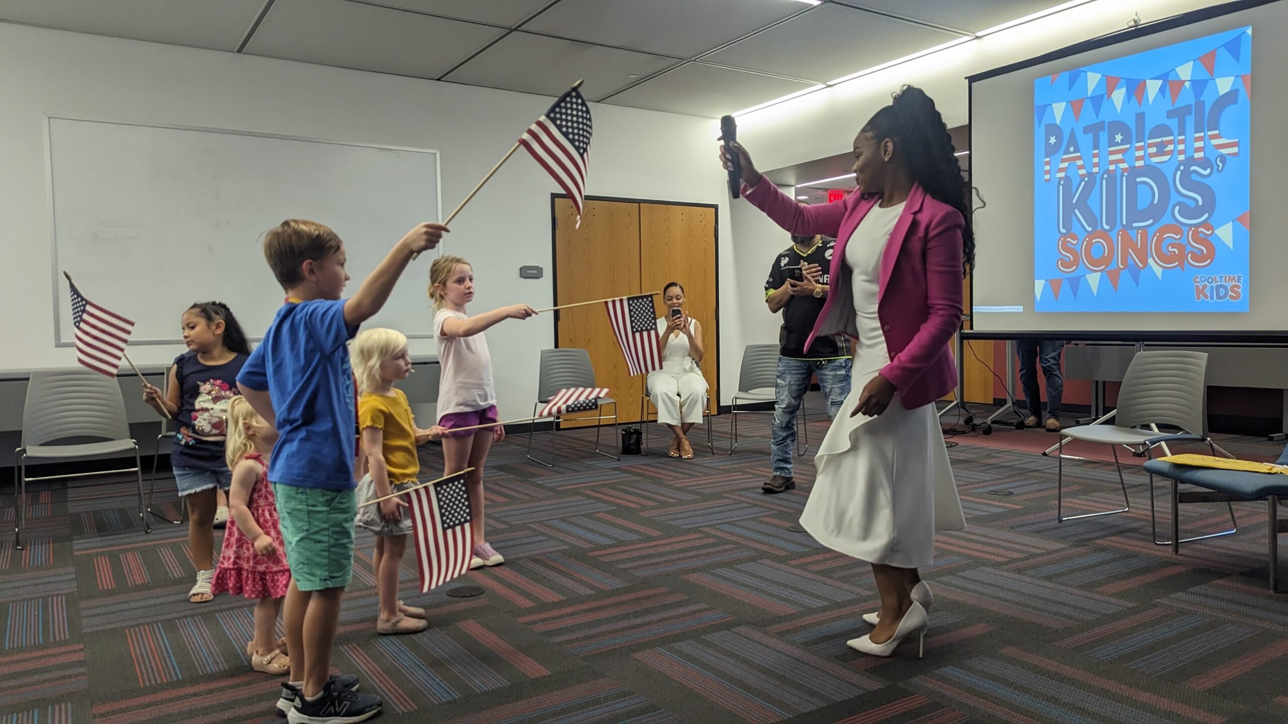 A few kids wave American flags under the direction of a Black woman at a Brave Books library event.