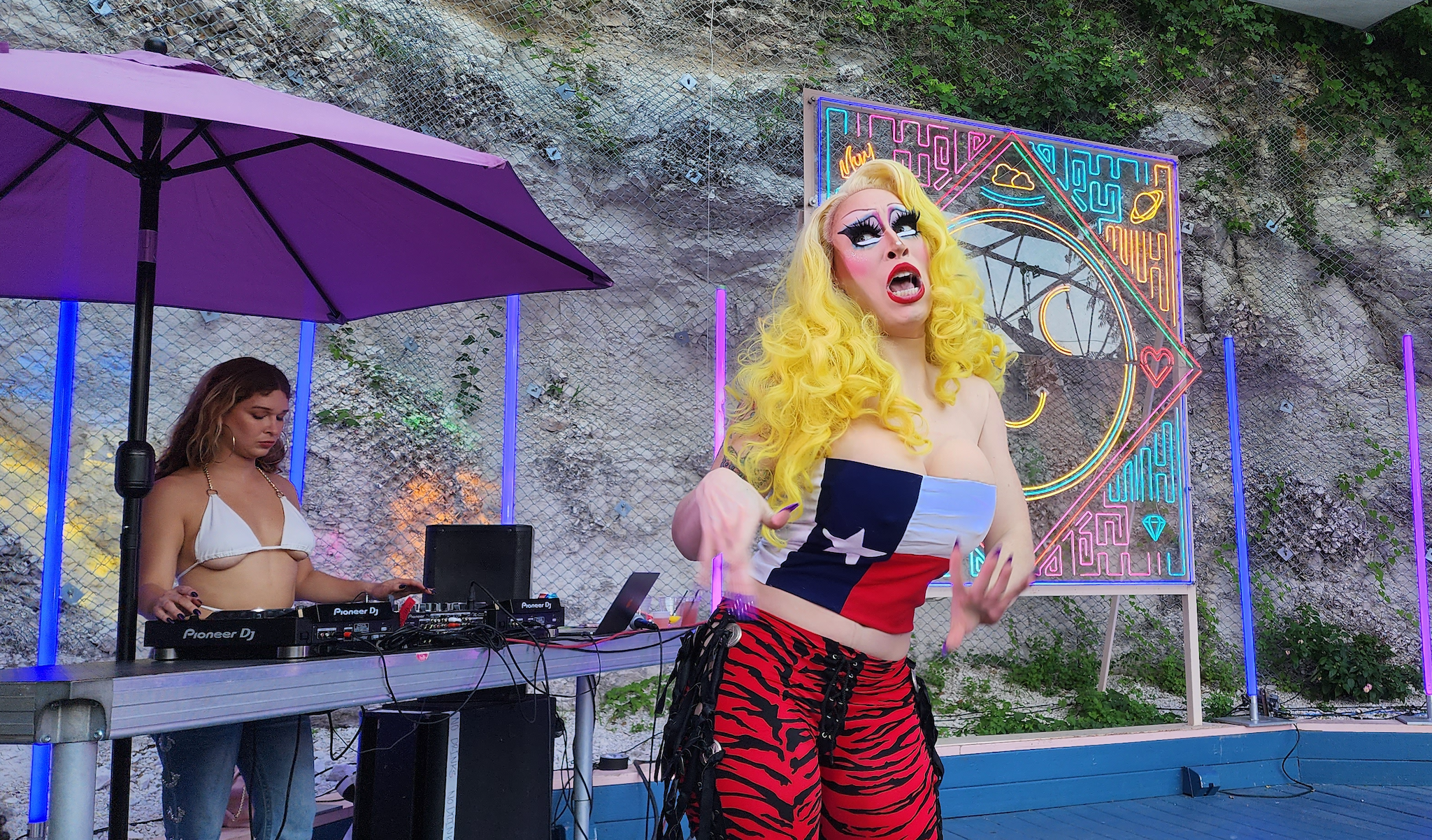 Drag queen Brigitte Bandit performs on an ouitdoor stage in a sleeveless Texas flag halter top over large prosthetic breasts, a bright yellow blonde wig, and red and black tiger-stripe side-tie cowboy pants.