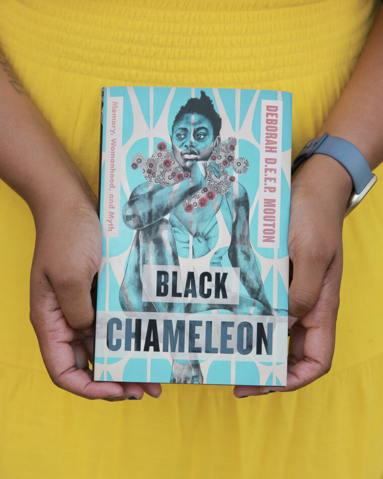 The arms of Deborah Mouton, a Black woman, seen in a yellow dress, holding her book Black Chameleon. She's got a tattoo on the underside of one arm and is wearing a blue watch band. cThe book's cover shows a Black girl with a flower-like pattern wreathing her neck.