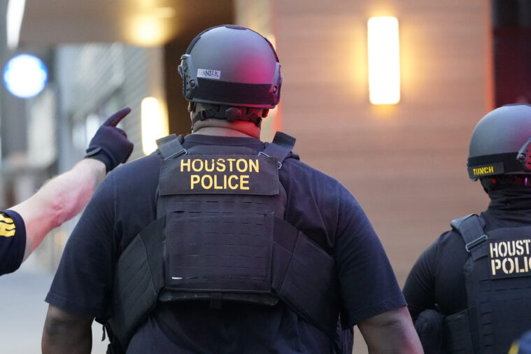 Three Houston police in riot helmets and armor, seen from behind. One, mostly off camera, is pointing the right.