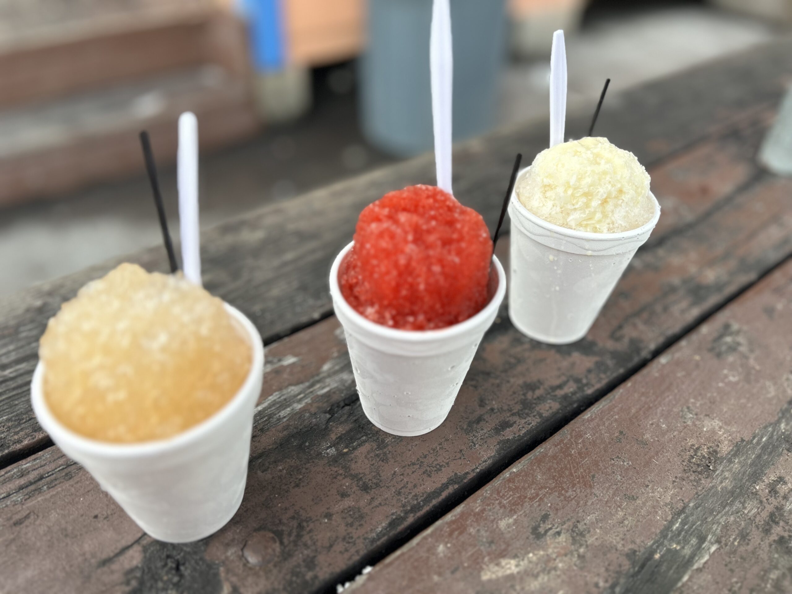 Three styrofoam cups of shaved ice with plastic spoons stuck in them, lined up in a row on a wooden countertop. The shaved ice is light orange, bright red, and light yellow.