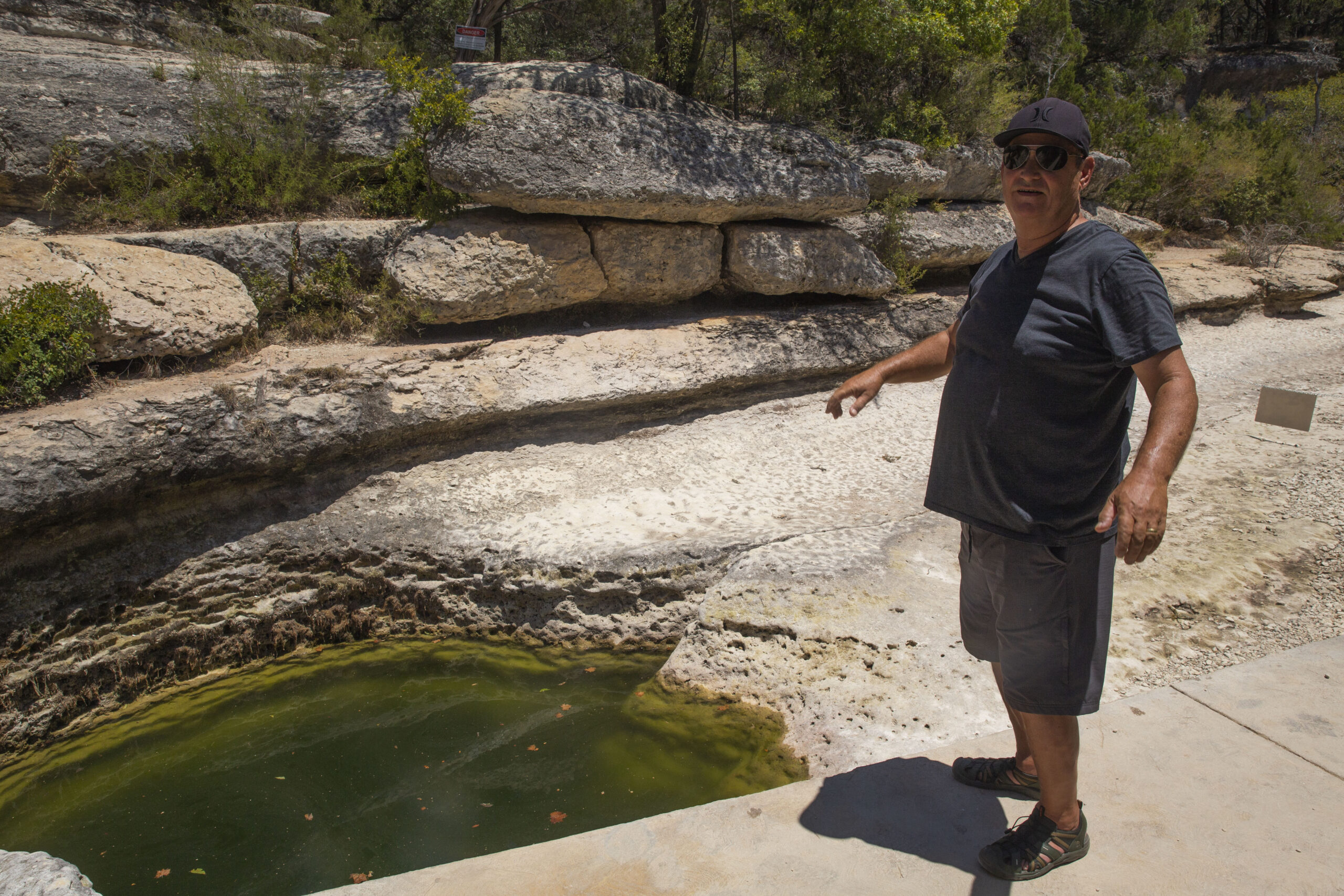 David Baker, a white man in blue shorts, t-shirt and ballcap points at the mucky green puddle where the famous spring known as Jacob's Well once flowed.