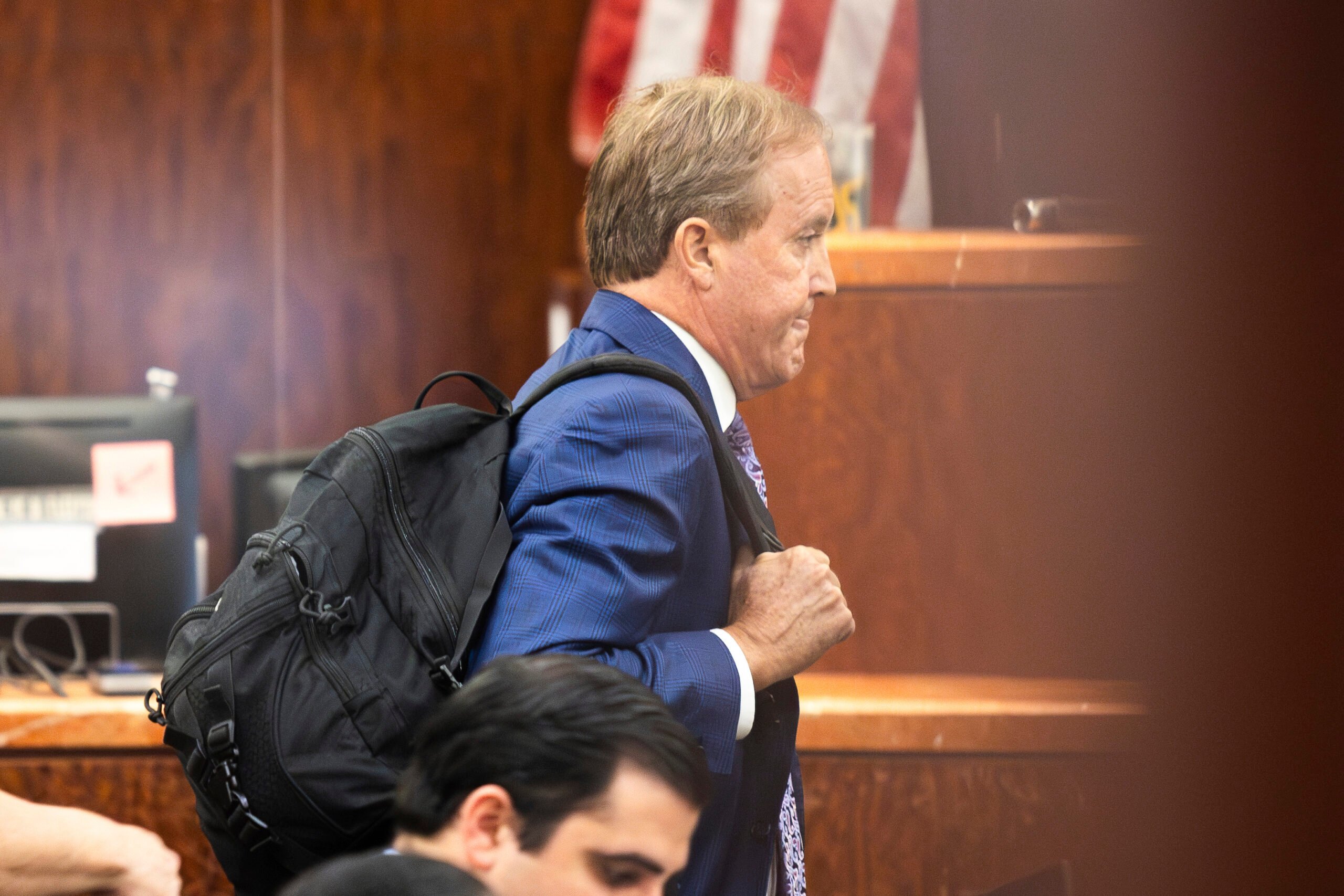 Ken Paxton, seen in profile in a blue suit jacket and black backpack, has a somewhat goofy, tongue-in-lips expression as he hoists his bag and carries it out of a courtroom.