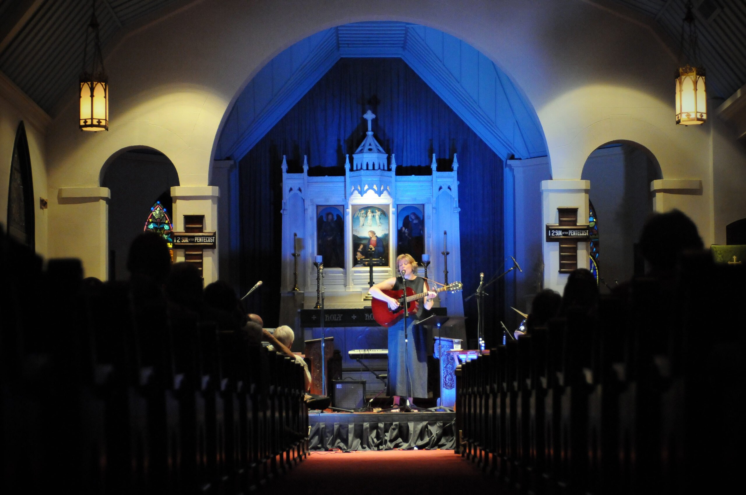 A performer plays music on the church Sanctuary, turned into a makeshift stage. The photo has a church-like atmosphere, with dimmed lights, viewers seated on pews and the altar behind the musicians.