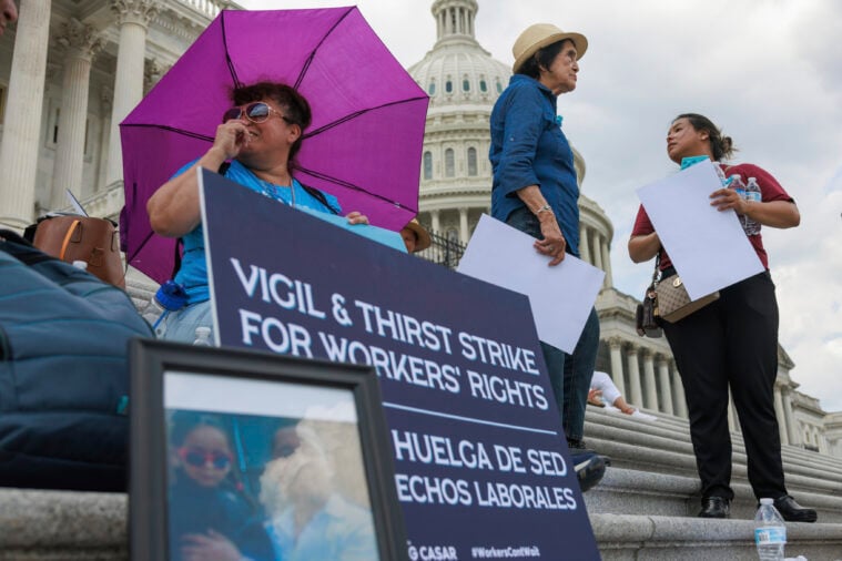 A worker sits on the steps of the U.S. Capitol in front of a small makeshift memorial and a sign advertising the "Vigil and Thirst Strike for Workers Rights." She is protecting herself from the sun under a pink umbrella.