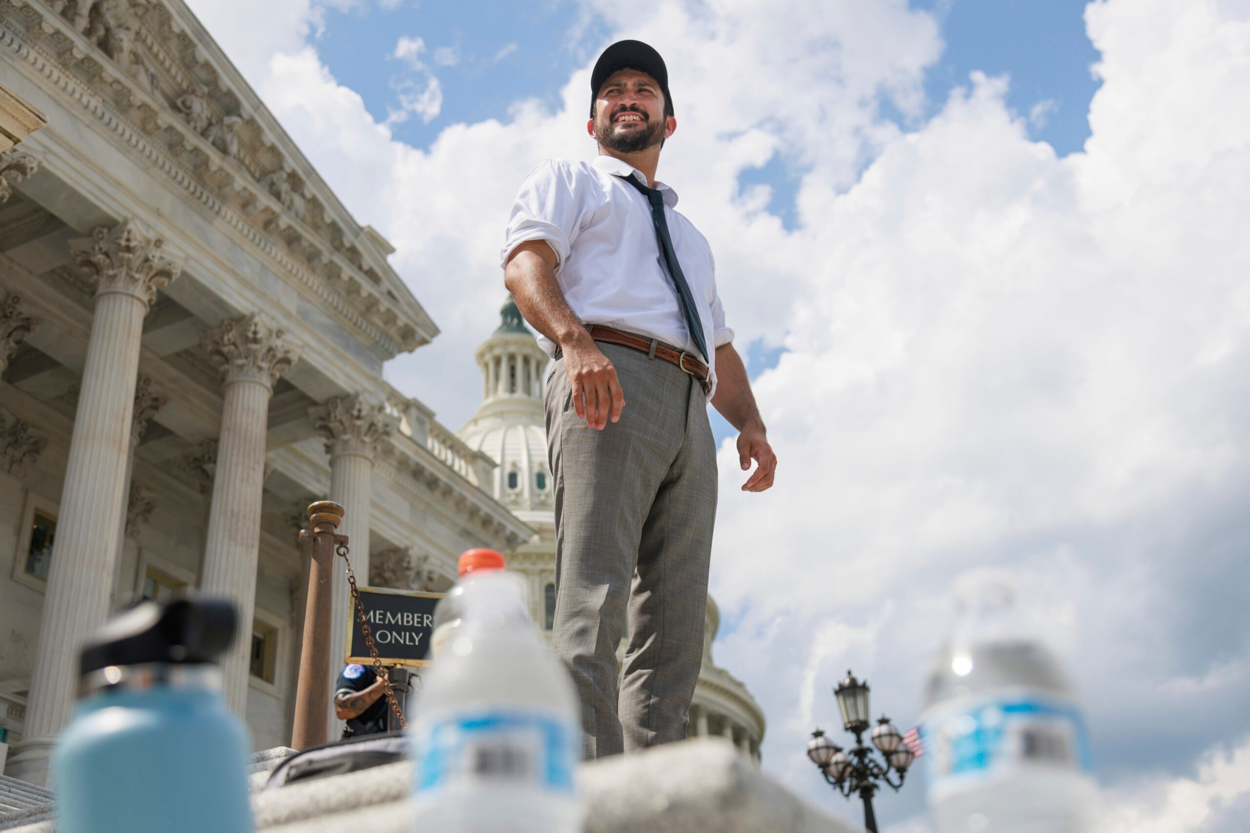 U.S. Representative Greg Casar (D-TX), a Latino man, is seen beyond water bottles on the steps of the U.S. Capitol in Washington, D.C. He's wearing formal pants, a shirt and tie, with his sleeves rolled up.