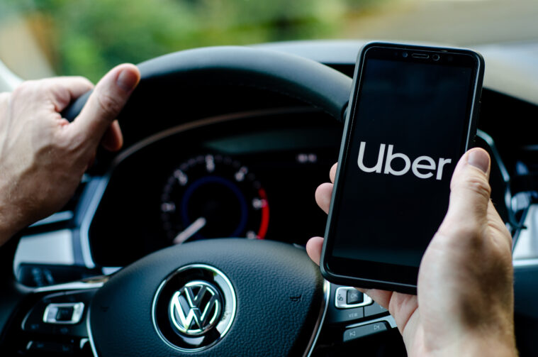 The hands of a driver in a fancy car. They hold their smarphone up, showing the Uber app logo on screen.