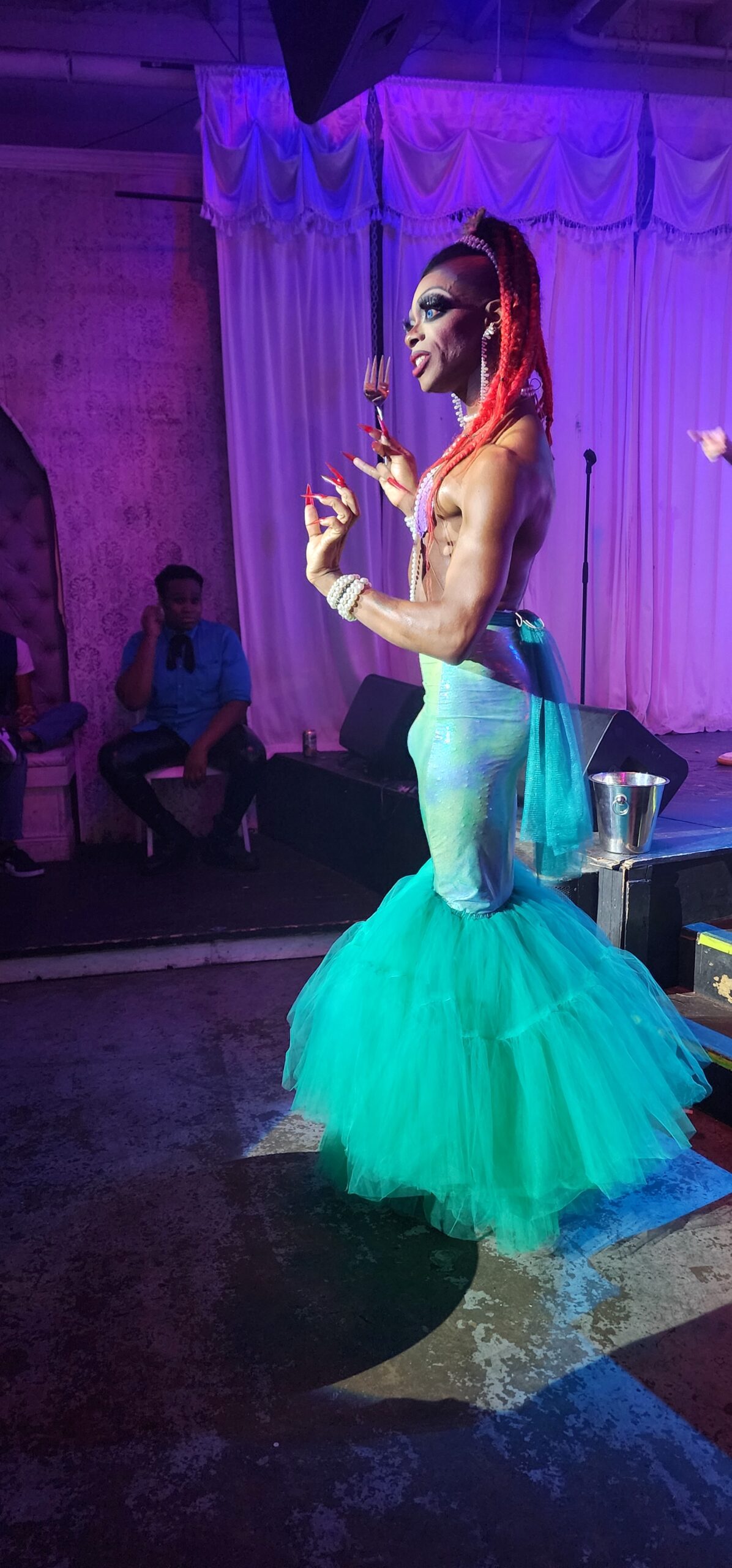 Hernajestie the Hung, a muscular, skinny Black drag queen in a sea green mermaid costume, makes an elegant gesture while holding an oversized prop fork. Her hair features bright red braids and shaved sides. 