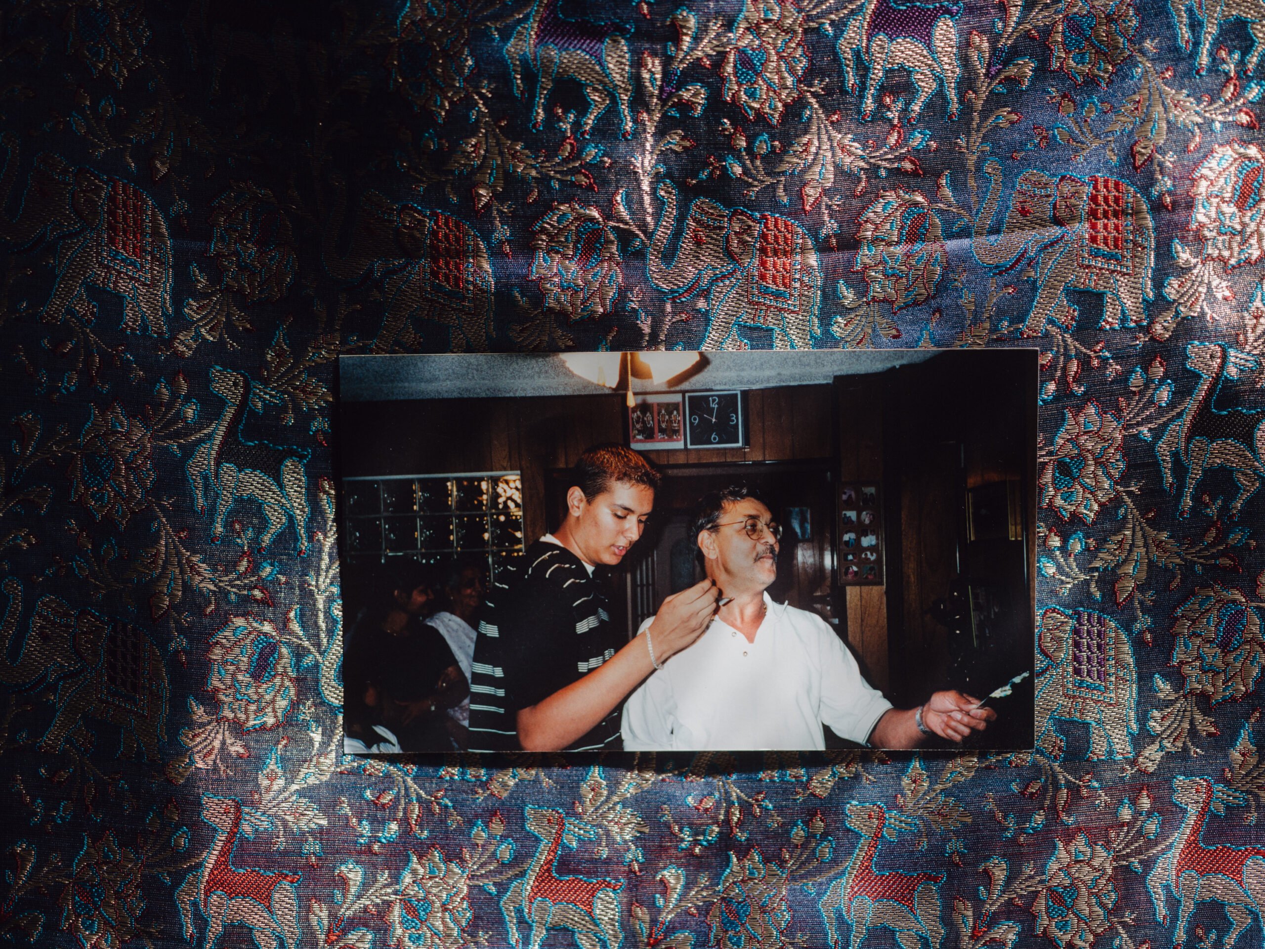 Against the backdrop of a blue sari fabric, a color photograph of a father and son, Hasmukh Patel and Mitesh Patel, posing at the father's 50th birthday.