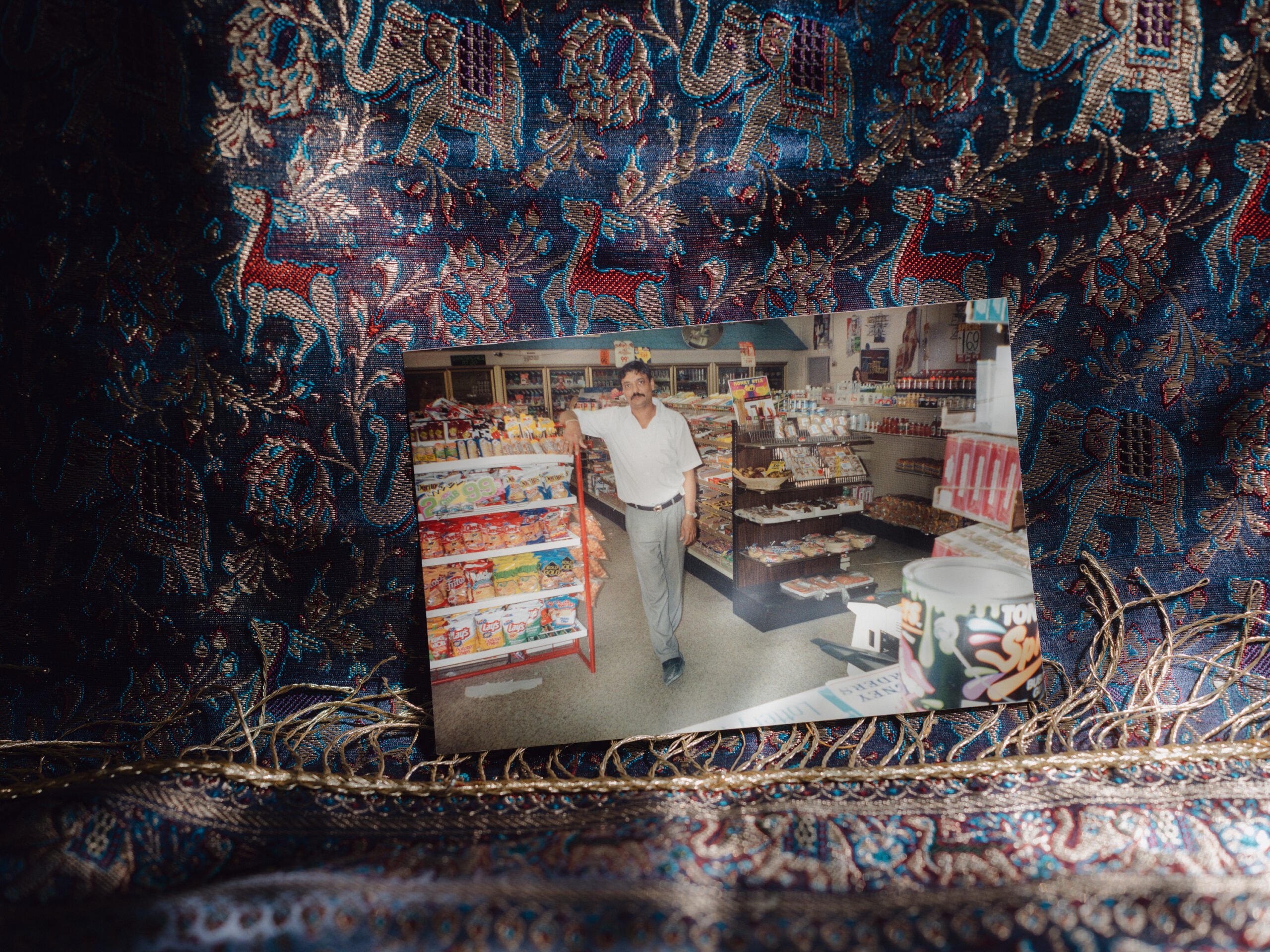 A photo of Hasmukh Patel in his convenience store is photographed on a backdrop of an old-fashioned Indian sari fabric printed with elephants and camels.