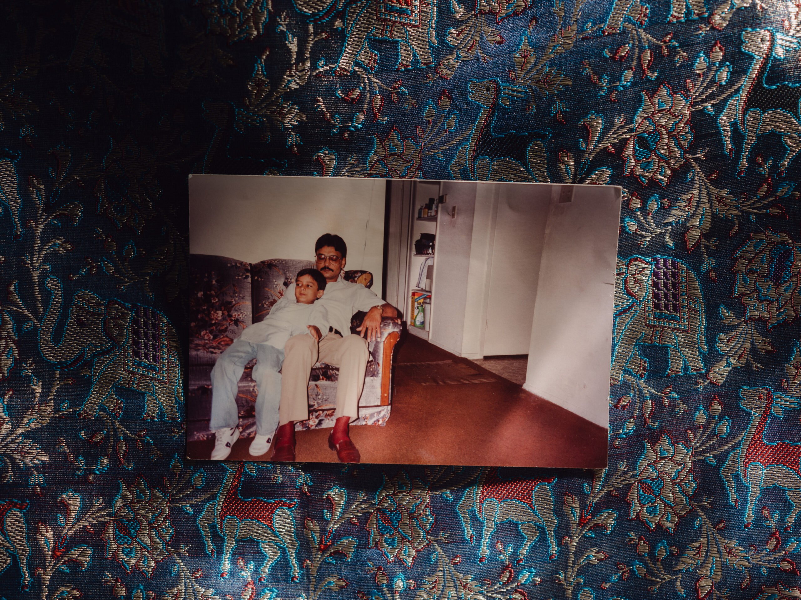 Hasmukh Patel lounges on the couch with his young son Mitel Patesh leaning against him, both wearing relaxed expressions and matching casual clothes.