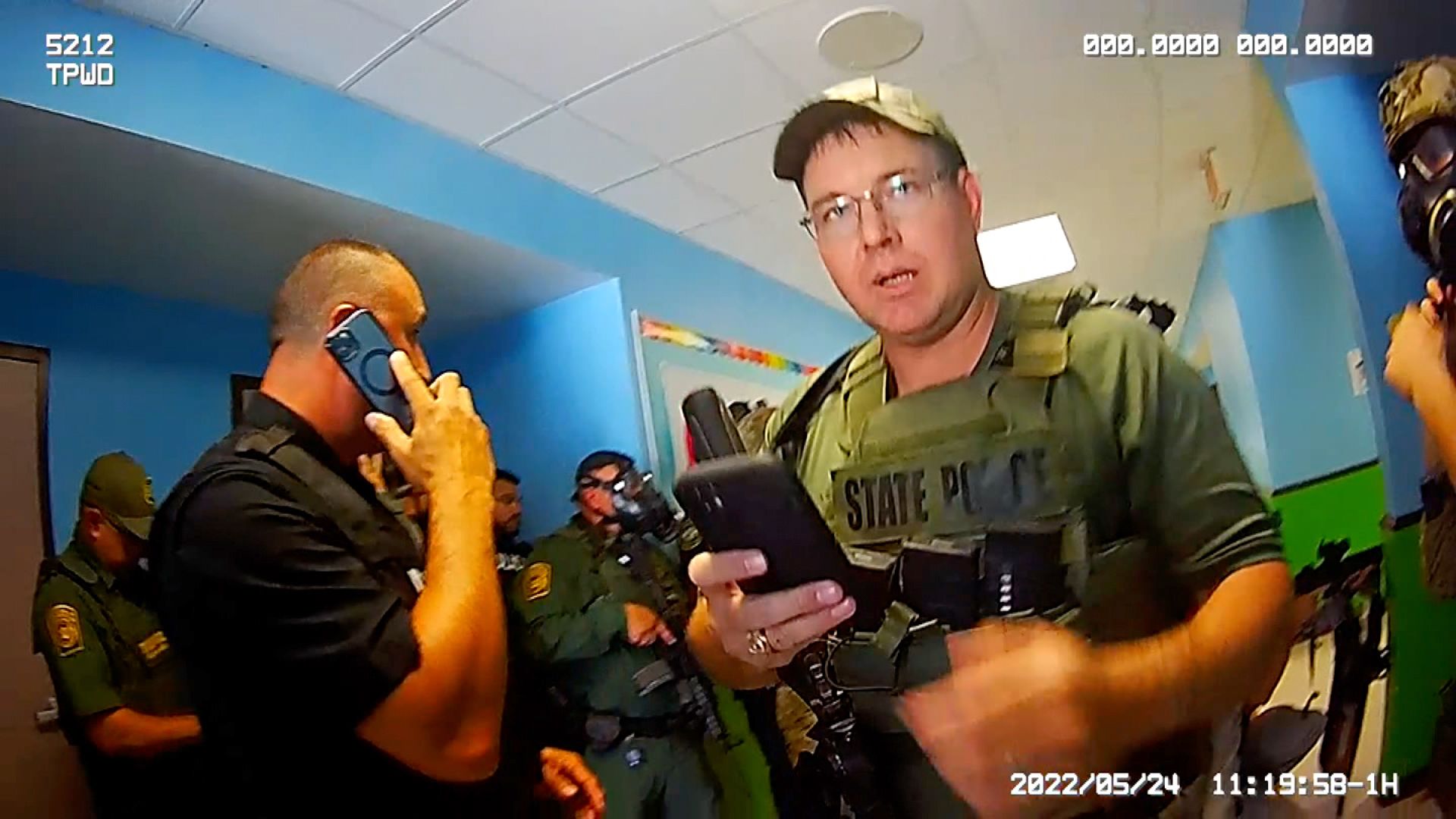 A photo shows body cam footage of Texas Ranger Ryan Kindell the day of the Uvalde massacre. Kindell looks at the camera while holding up his phone. Officers in the background are also on their phones. 