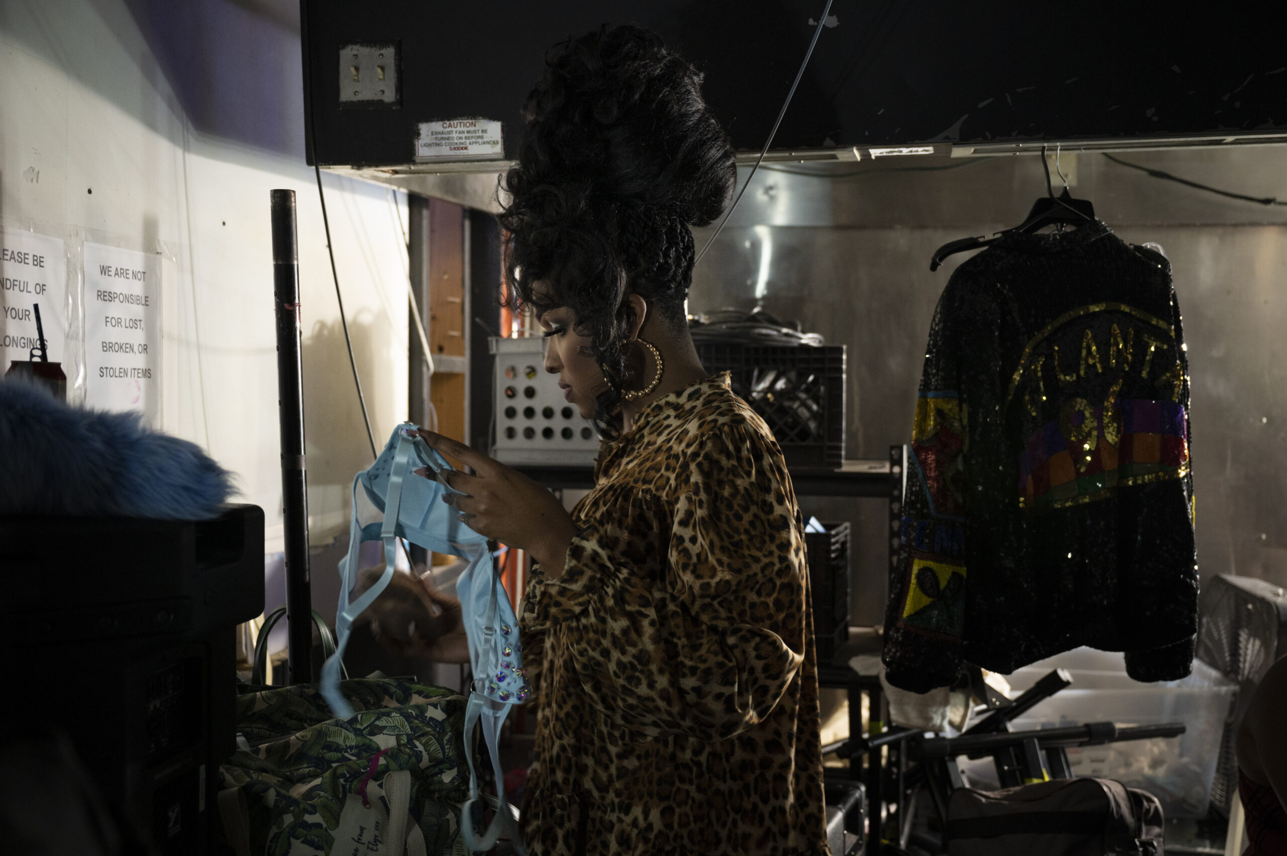 A Black woman with a very tall up-do and big hoop earrings, wearing a leopard print coat. She's holding a blue costume bra top in her hands as she stands in a backstage area.