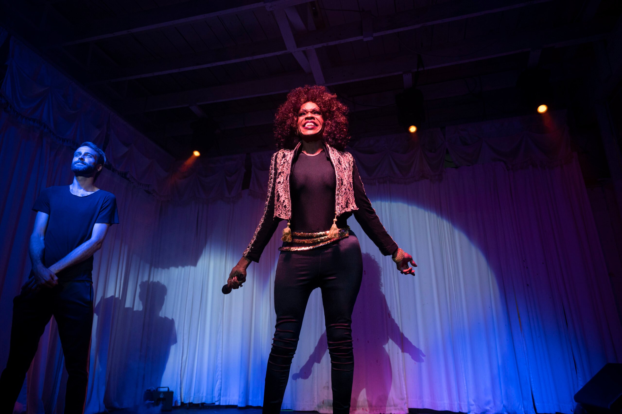 Ray Ray Topaz, an older Black drag queen who looks like Tina Turner, performs in a black outfit with pink and black jacket, standing with her arms spread wide behind her proudly, the microphone in one hand. The sign language interpreter stands to one side.