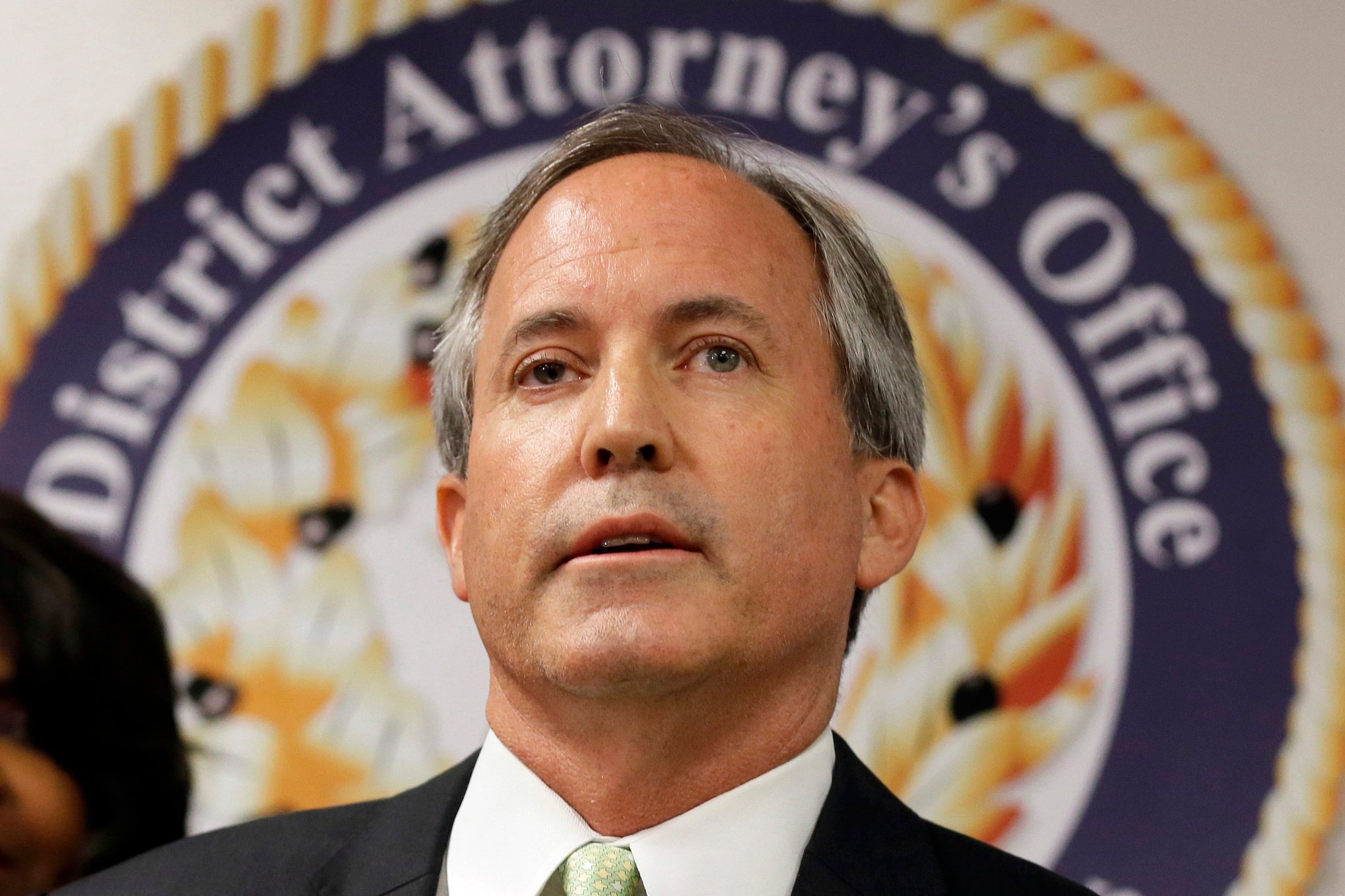 A close-up portrait of Attorney General Ken Paxton, in a suit and tie, standing in front of the seal of his office.