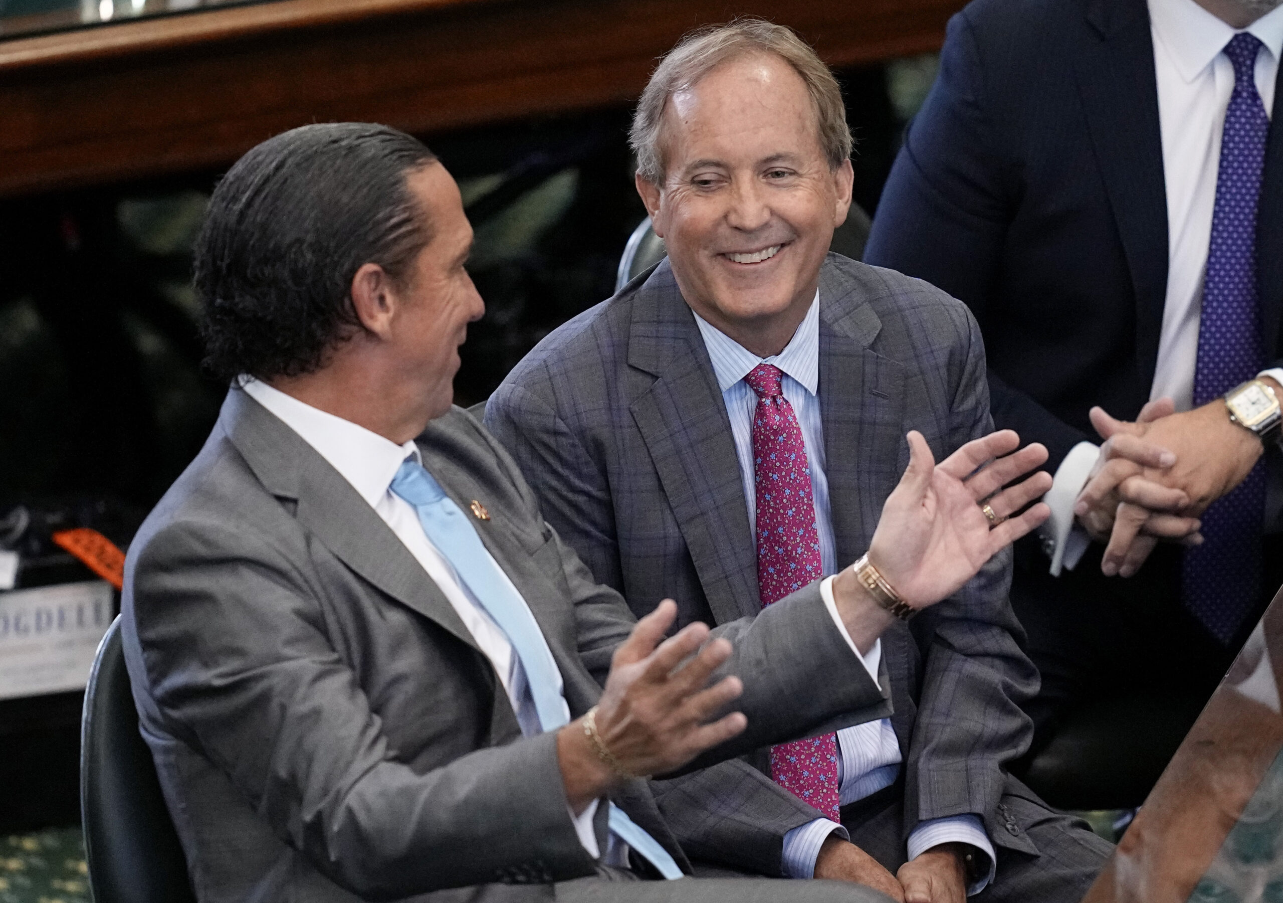 Ken Paxton and his lawyer are both wearing suits and ties, seated in the Senate chambers just before he was acquitted. Paxton's lawer, Tony Buzbee is gesturing with his hands while Paxton looks on with a grin.