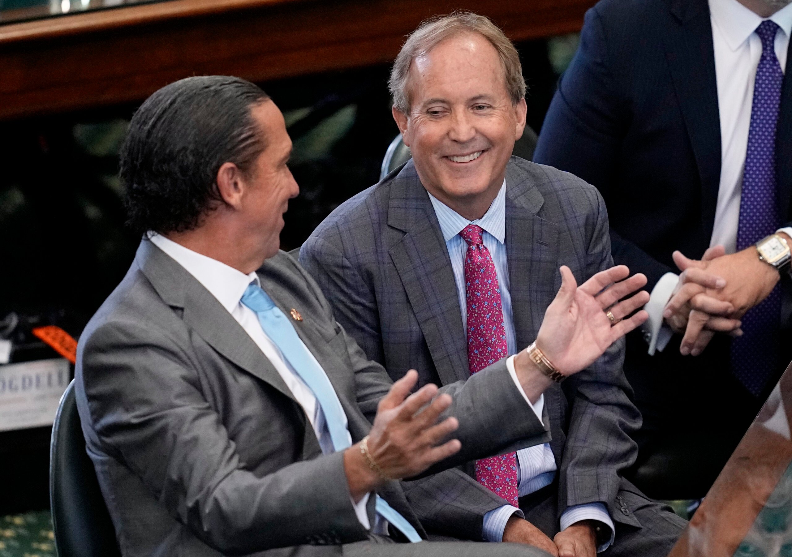 Ken Paxton and his lawyer are both wearing suits and ties, seated in the Senate chambers just before he was acquitted. Paxton's lawer, Tony Buzbee is gesturing with his hands while Paxton looks on with a grin.