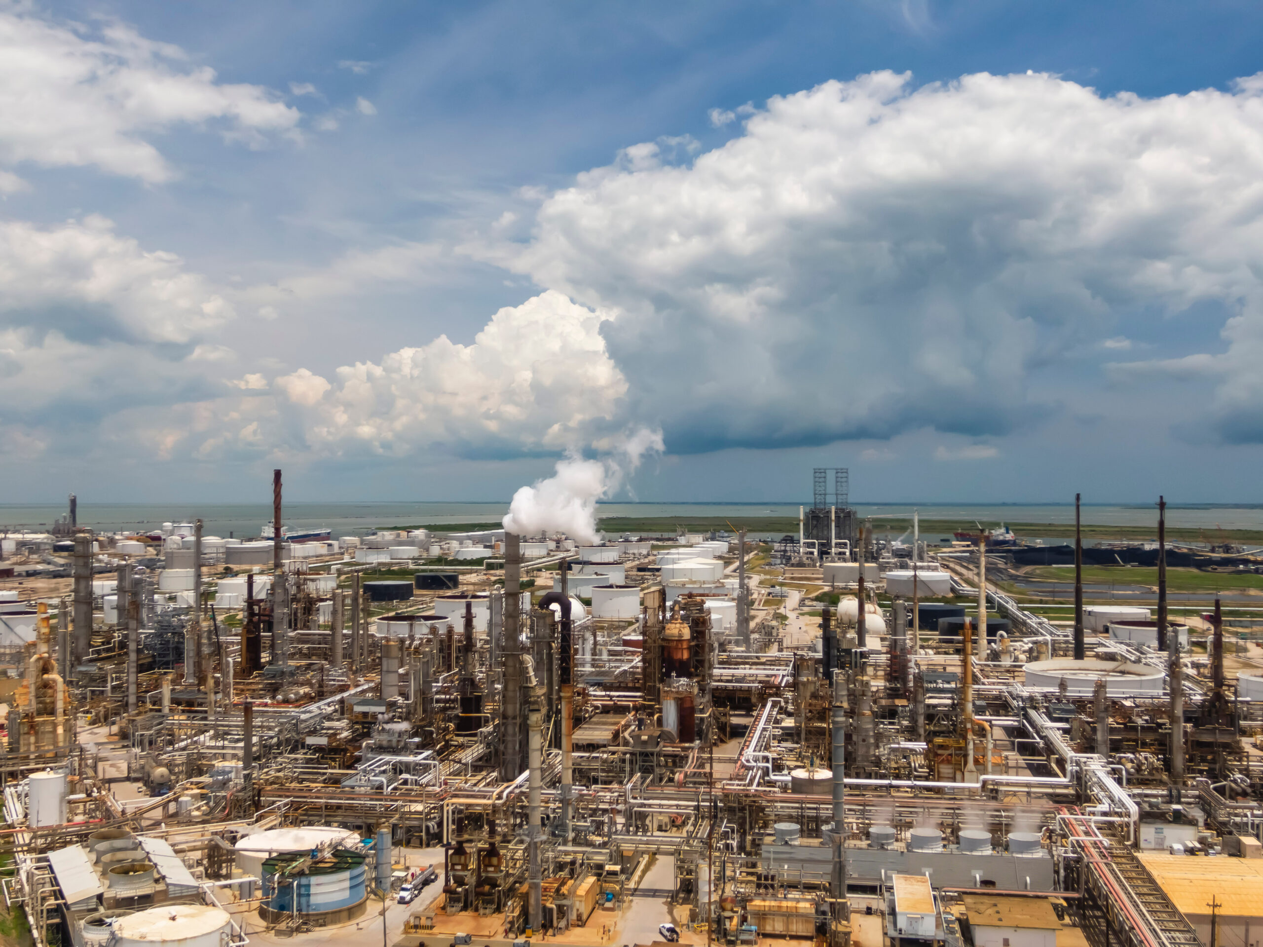 An aerial view of an oil and gas refinery on the Gulf Coast in Texas City. Smokestacks and storage containers dot the landscape under a partly cloudy sky.