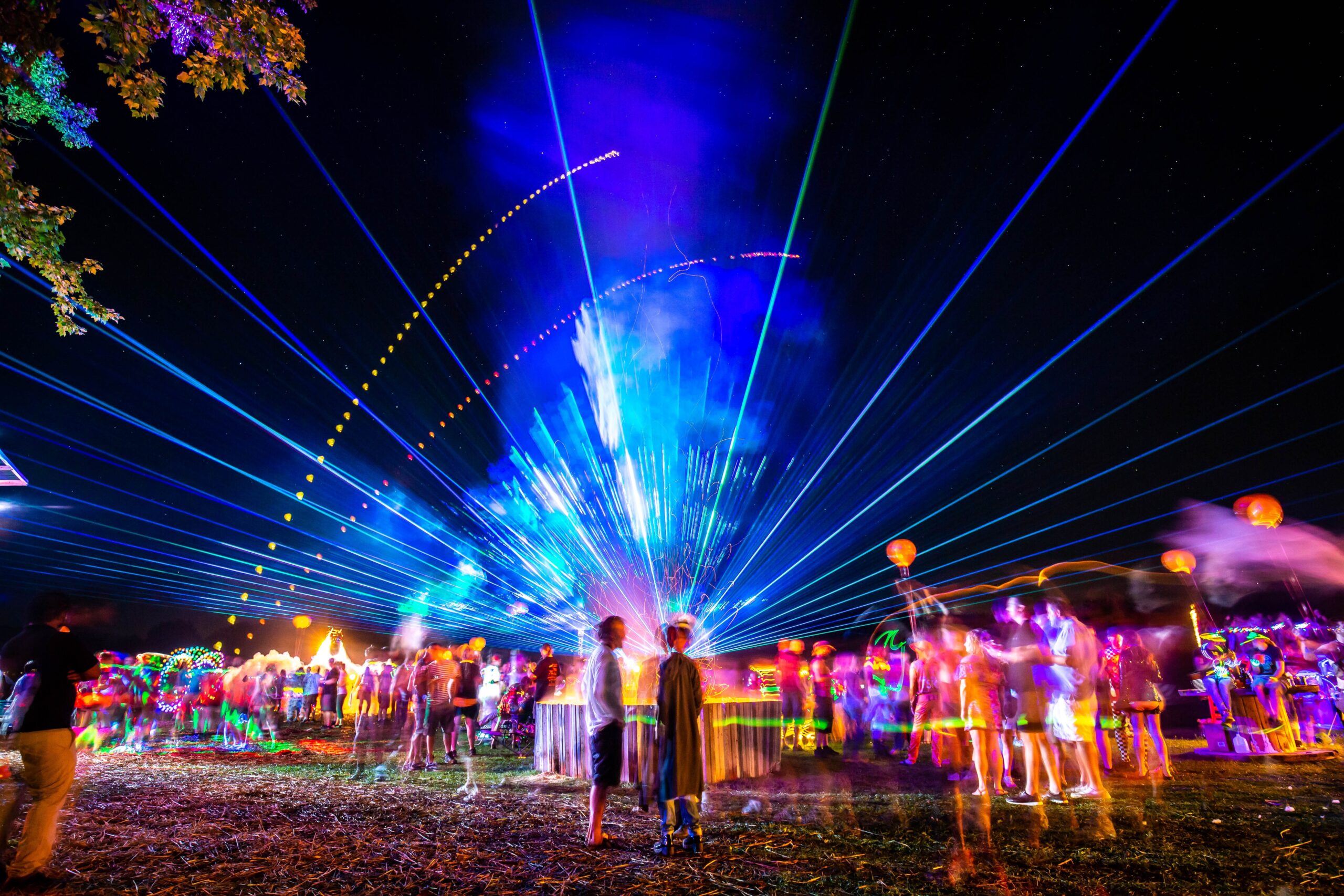 An artistically blurred photo of an independent or underground music party at night, with colorful lights and lasers leaking into the sky, as people cluster about in groups enjoying themselves.