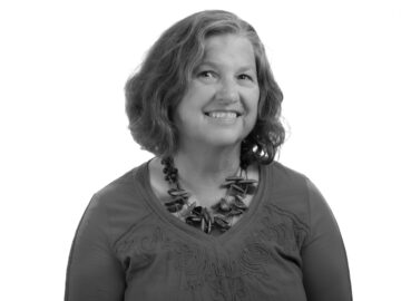 A black and white portrait of Paula Levihn-Coon, a white woman with shoulder length brown hair, in a sweater and chunky beaded necklace.