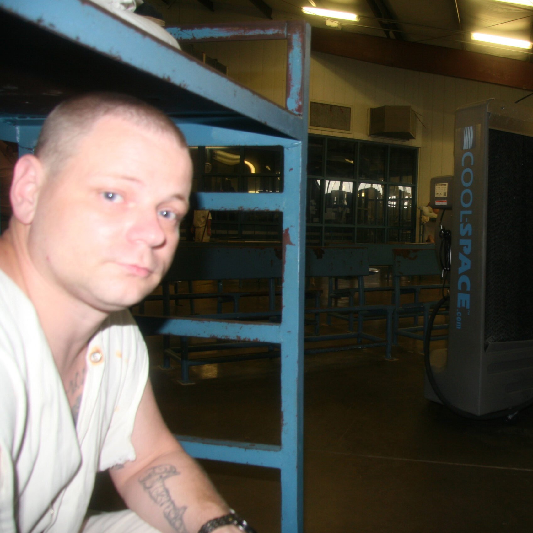 A prisoner sits on his bunk in his white prison uniform, looking at the camera. The feet and legs of another prisoner sitting on an upper bunk is also visible. A cooling fan is installed nearby.