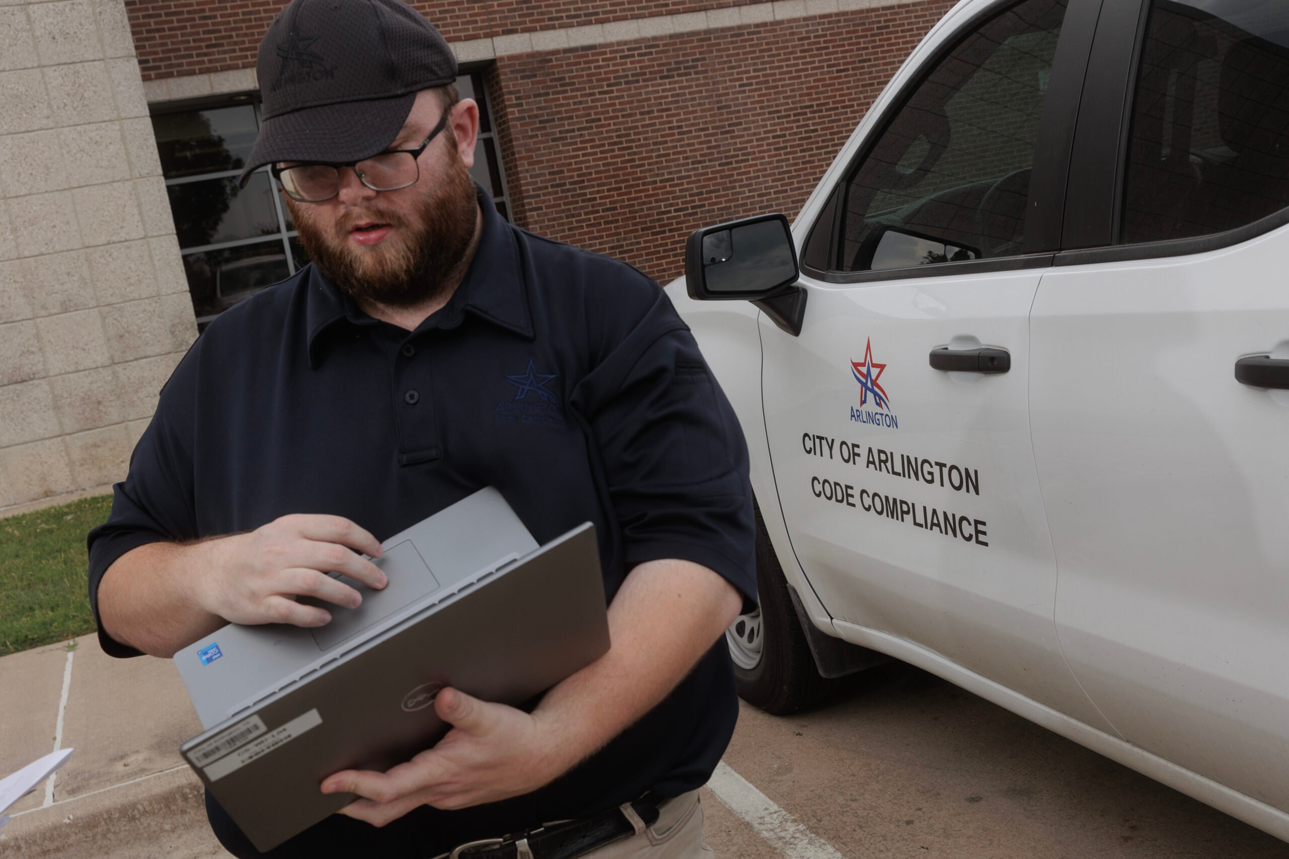 A thickly bearded man in a black cap and black polo scrolls through a laptop held in his otherh and, while standing by a City of Arlington Code Compliance enforcement vehicle.