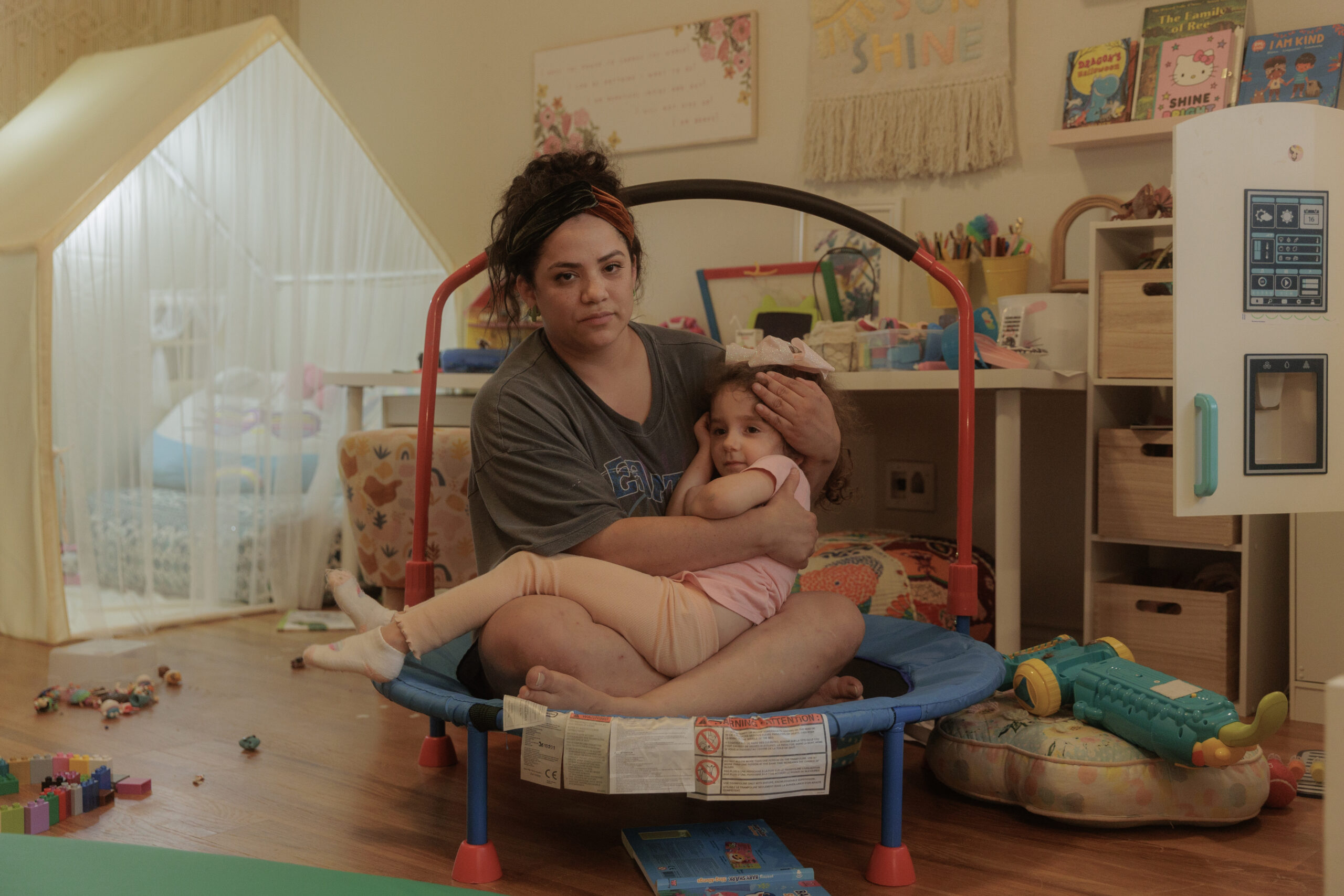 A Latinx woman cradles her young toddler-aged daughter in her lap, both wearing a concerned expression. A typical playroom, with scattered toys and a play tent, form the photo's backdrop.