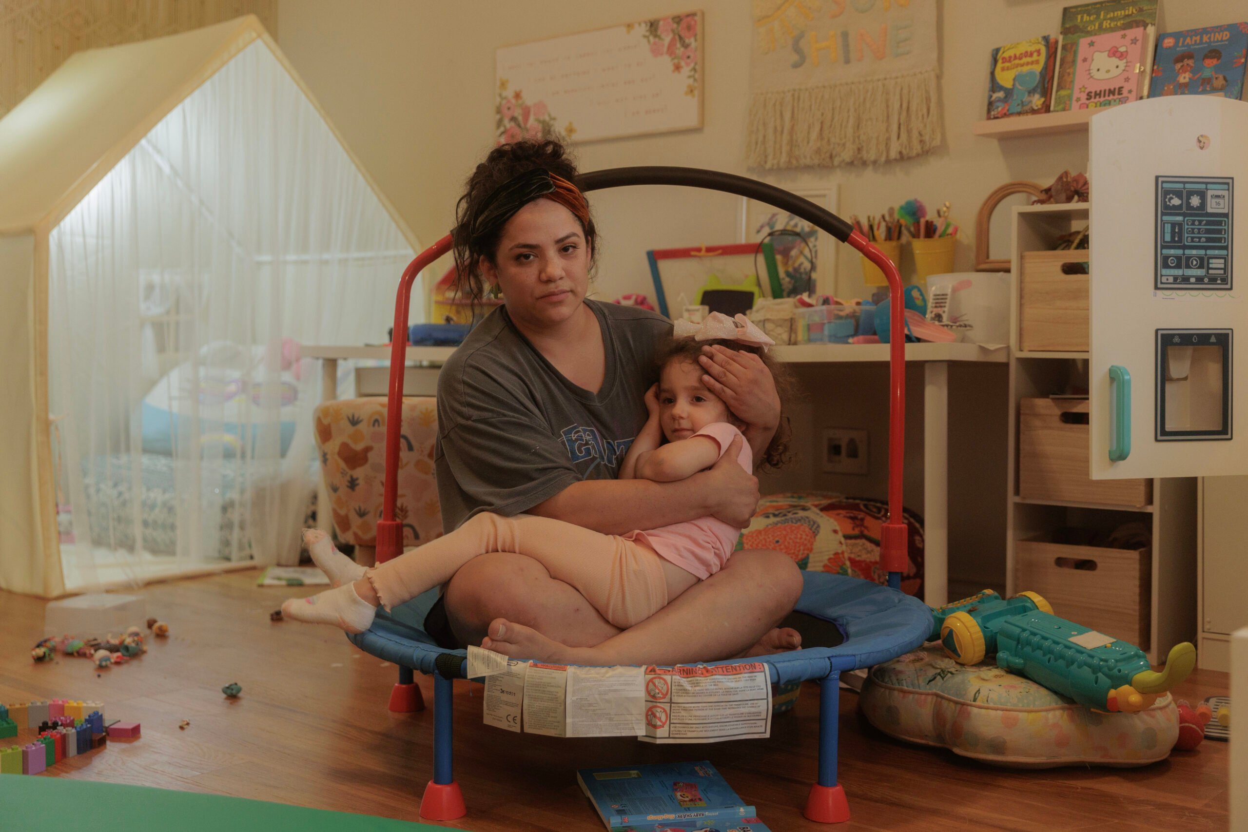 A Latinx woman cradles her young toddler-aged daughter in her lap, both wearing a concerned expression. A typical playroom, with scattered toys and a play tent, form the photo's backdrop.