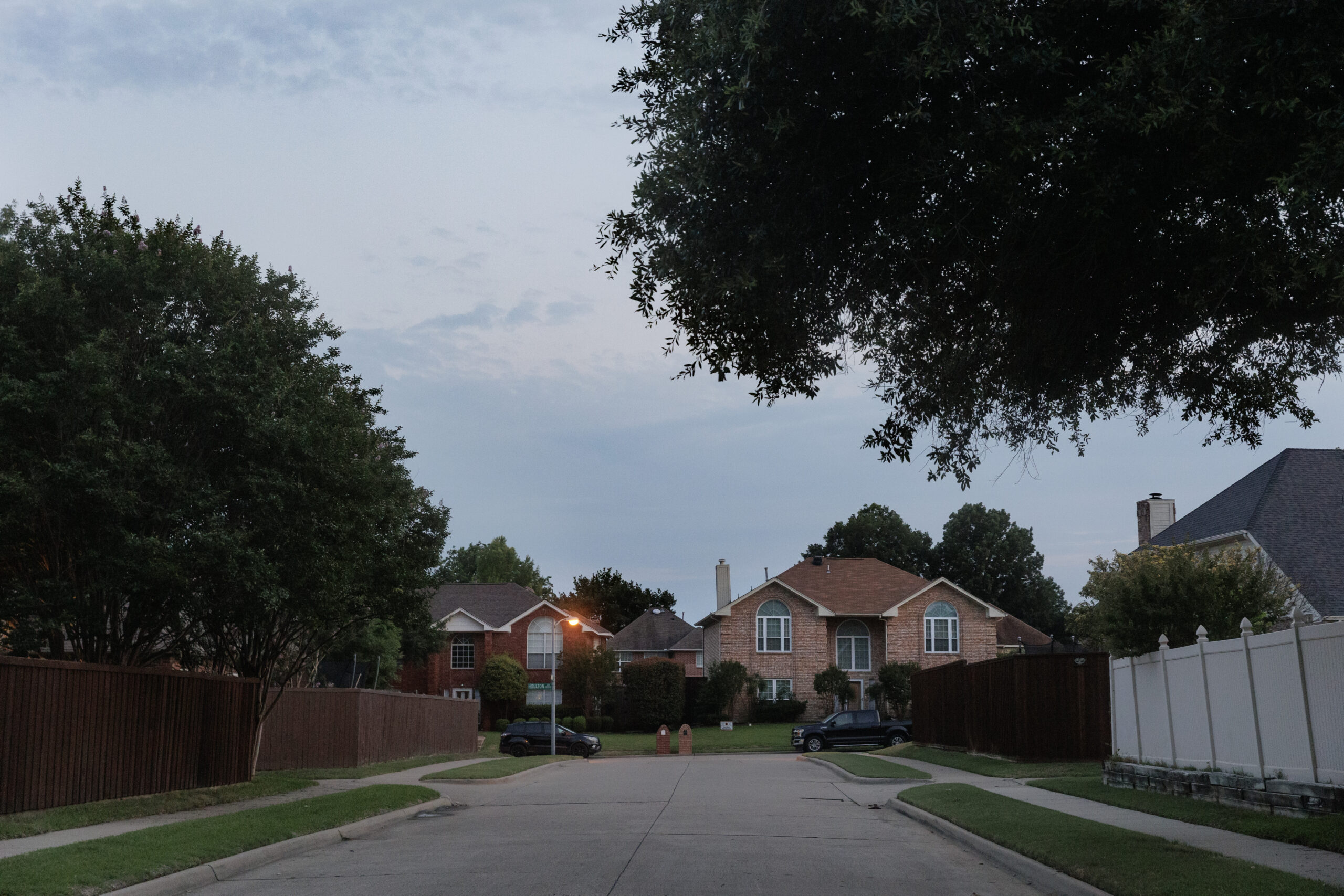 A tree-lined neighborhood with multi-bedroom, residential homes at dusk, the street lights beginning to turn on.