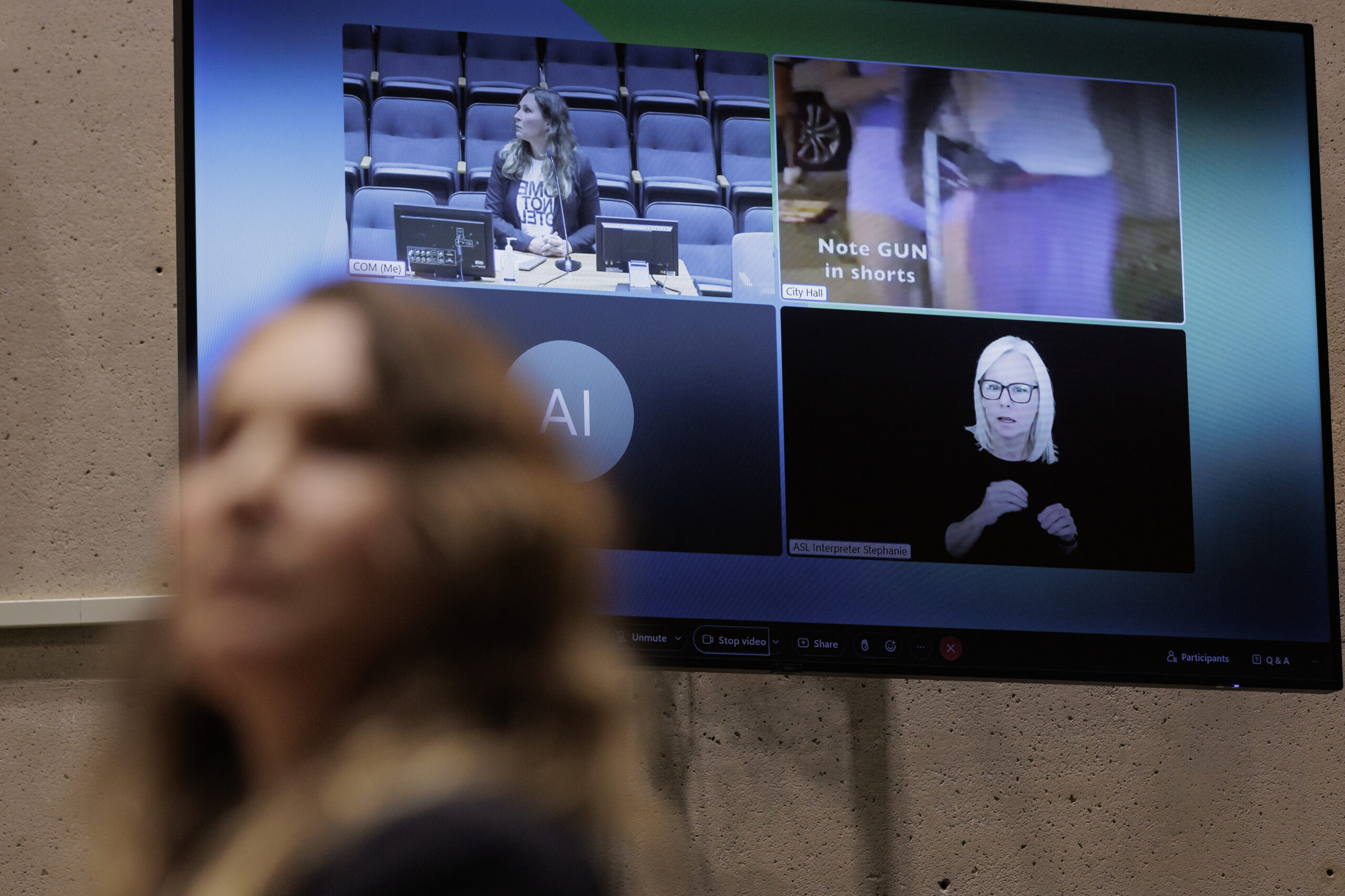 A brown-haired woman is out of focus in the foreground, while the camera clearly shows the video screen behind her, which is showing footage of her, city council members, and a still from her video which shows a man in shorts, with a label pointing out the visible bulge of a gun in his pocket.