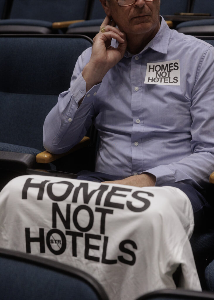 A man wearing a blue button down sits in a theater-style chair, wearing a Homes Not Hotels sticker with a similar t-shirt draped over the seat row in front of him.