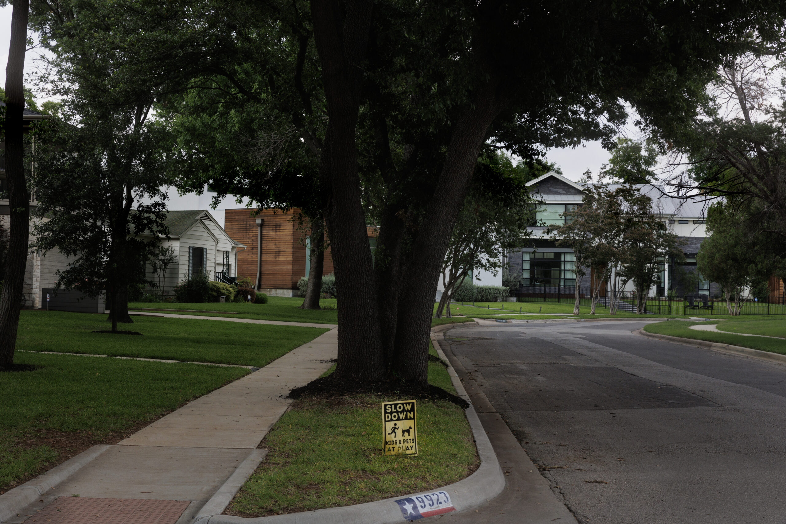 A residential street with modern mid- to large-sized homes, and a huge oak growing in the foreground. At the base of an oak is a yellow sign reading "Slow down, Kids & Pets at Play." The street looks placid. No cars are currently visible.