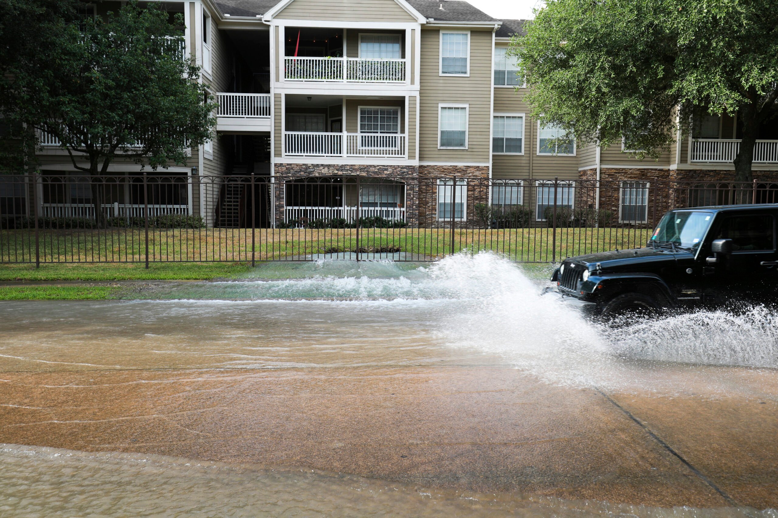 A dark-colored SUV plows into a massive puddle of water, spewing from broken pipes at the sidewalk near an urban Texas apartment building in Houston, one of many severe water leaks requiring repairs during the heatwave.