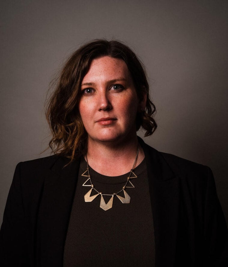 Loren Lynch is a white woman with a serious expression, wavy brown hair that's parted to her right side. She's wearing a black jacket, black shirt, and a gold necklace with angular brass pendants on it.