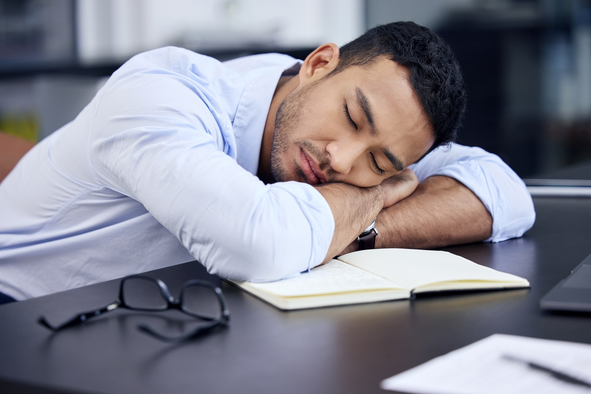 A POC worker lays his head down on his desk, closing his eyes in a nap. A pair of glasses and an open book are nearby, along with a notebook and pen.