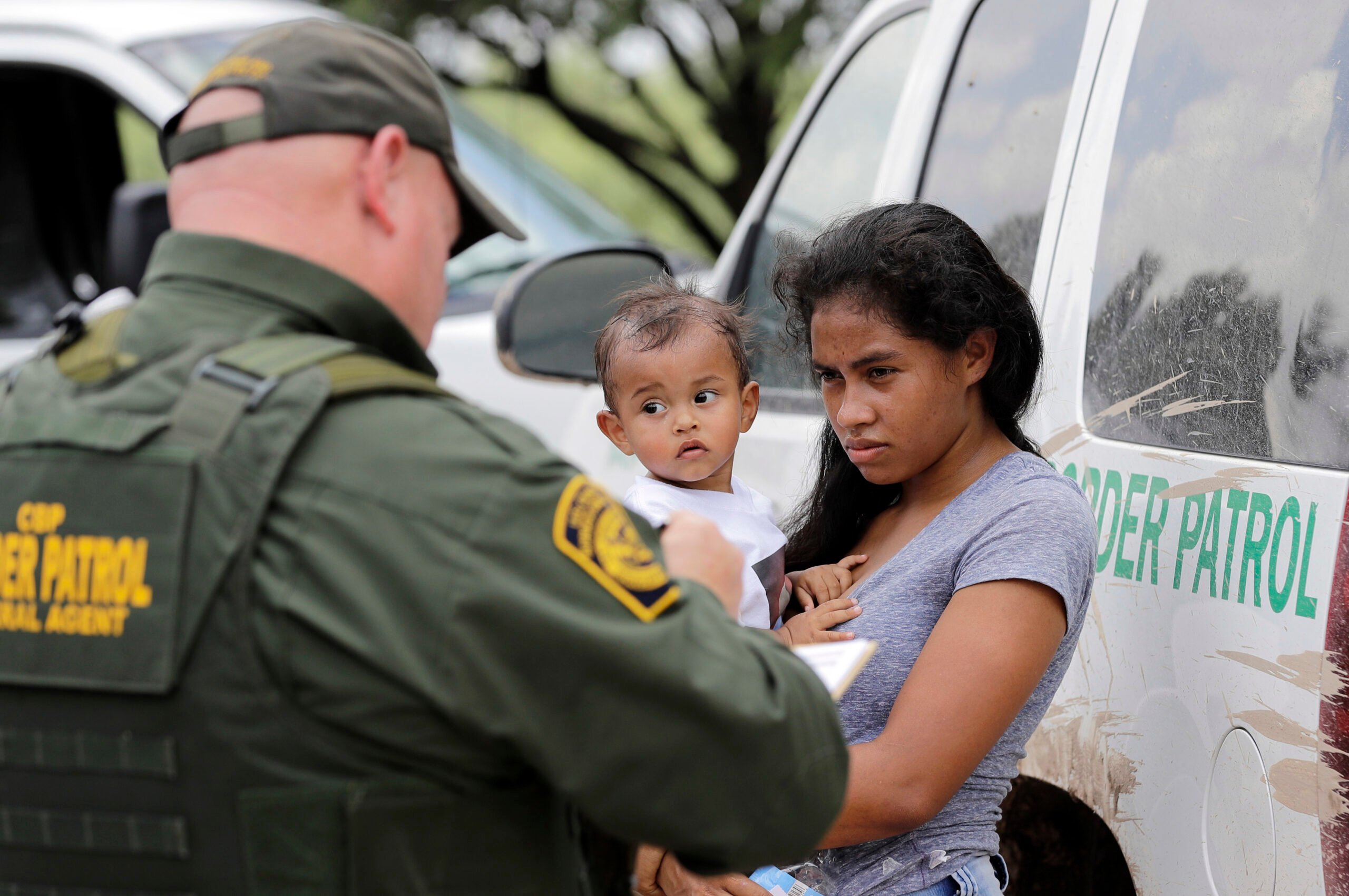 A border patrol officer in a green uniform interviews a Latina migrant holding a baby, while both are standing in front of a U.S. Customs and Border Patrol van.